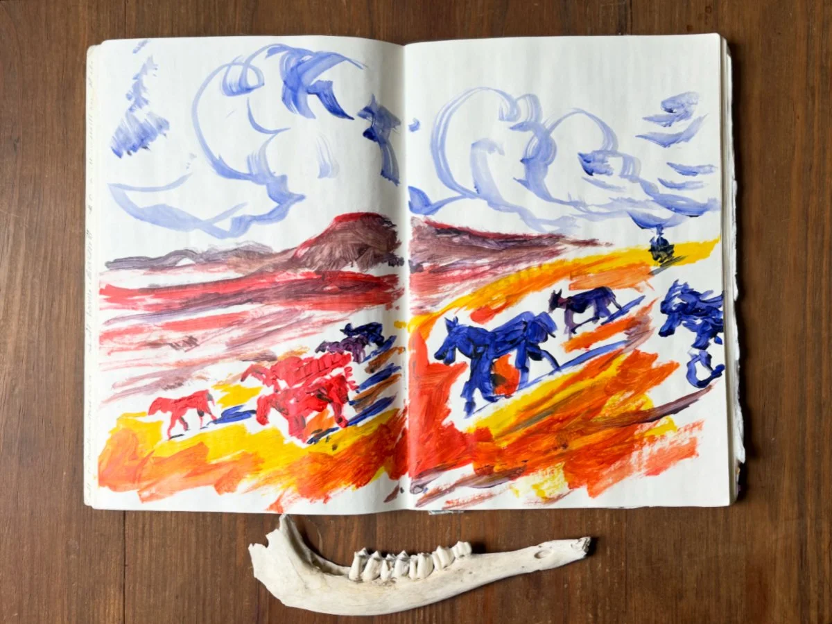 Open sketchbook acrylic painting of running horses beneath swirling clouds, beside a jawbone on a wooden table