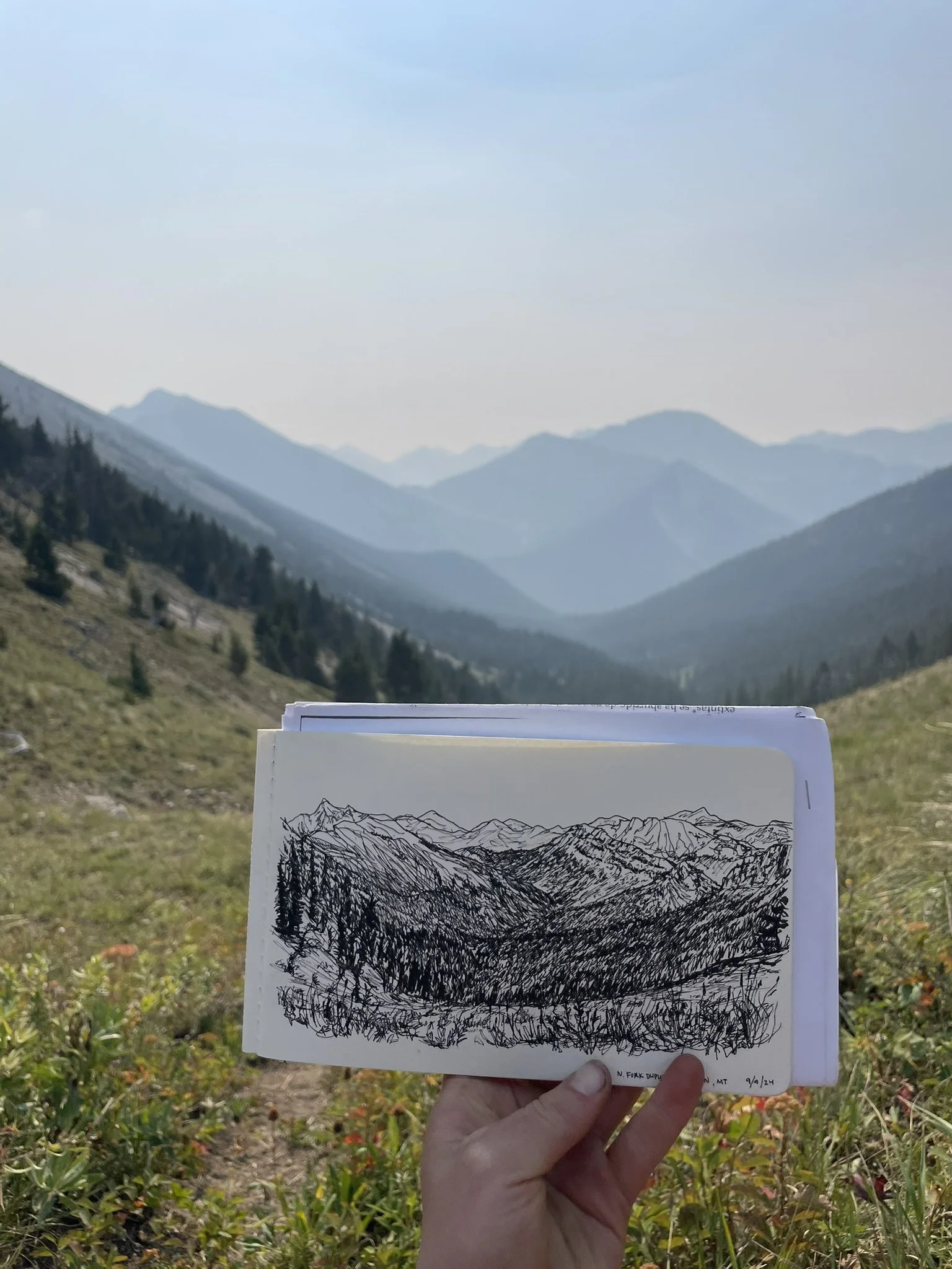 Artist holds a detailed mountain landscape sketch against the alpine valley and forested peaks it depicts.