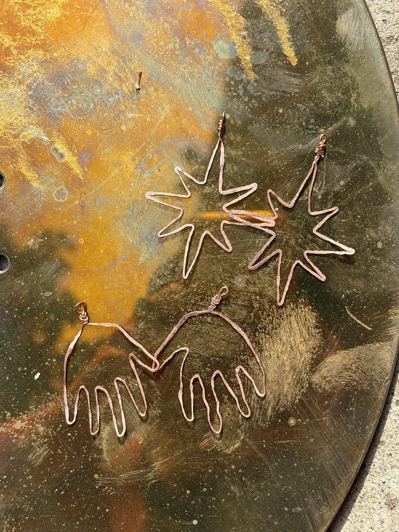 Handcrafted hammered copper wire wire sunburst and hand earrings on a weathered, bronze-colored metal surface.