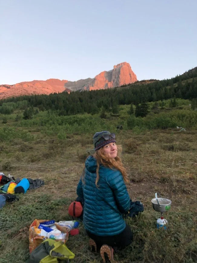 Hiker in a teal jacket kneeling at a campsite beneath red rock mountains at sunset
