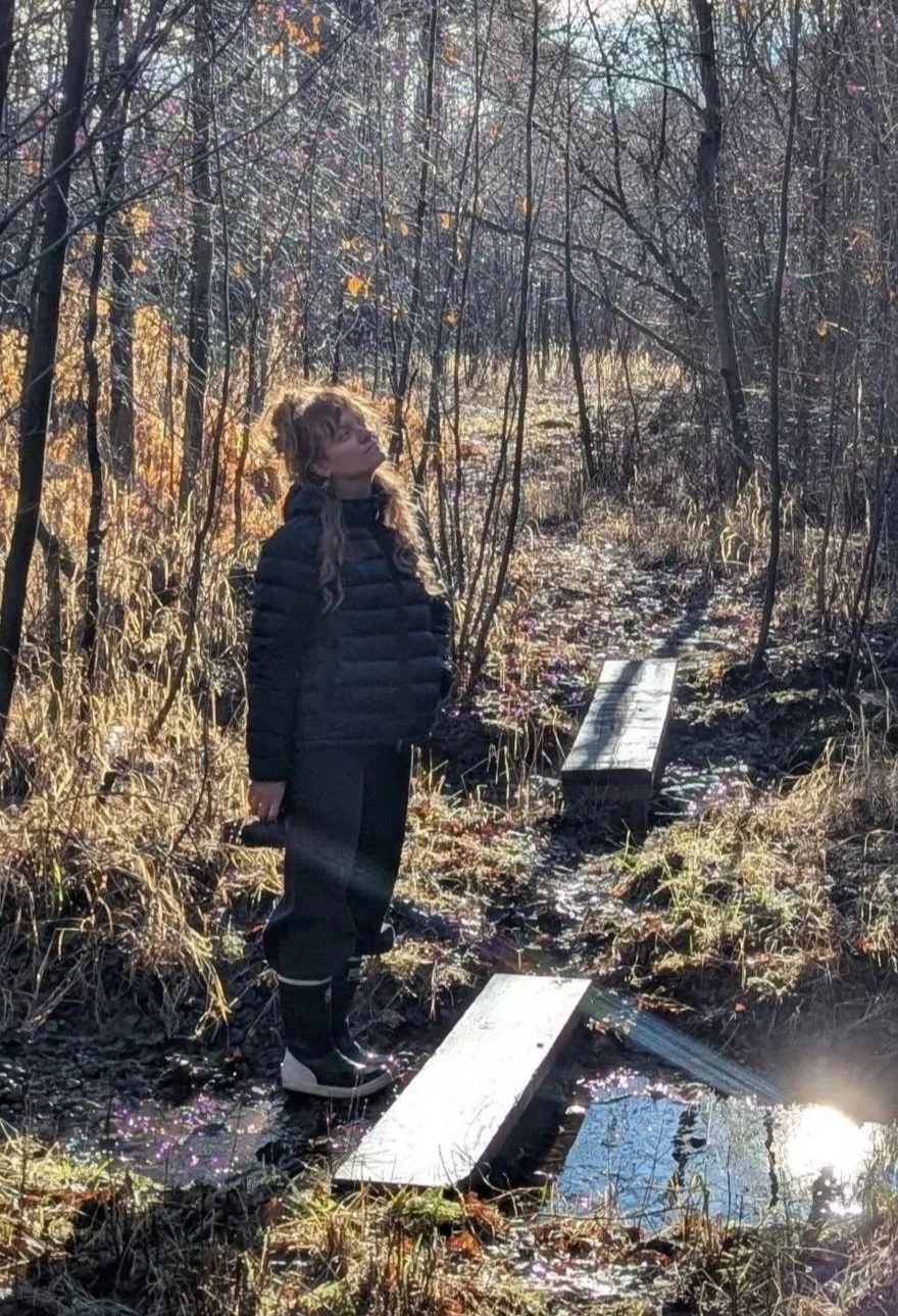 Person in a black puffer jacket crossing a narrow wooden footbridge over a forest stream in autumn