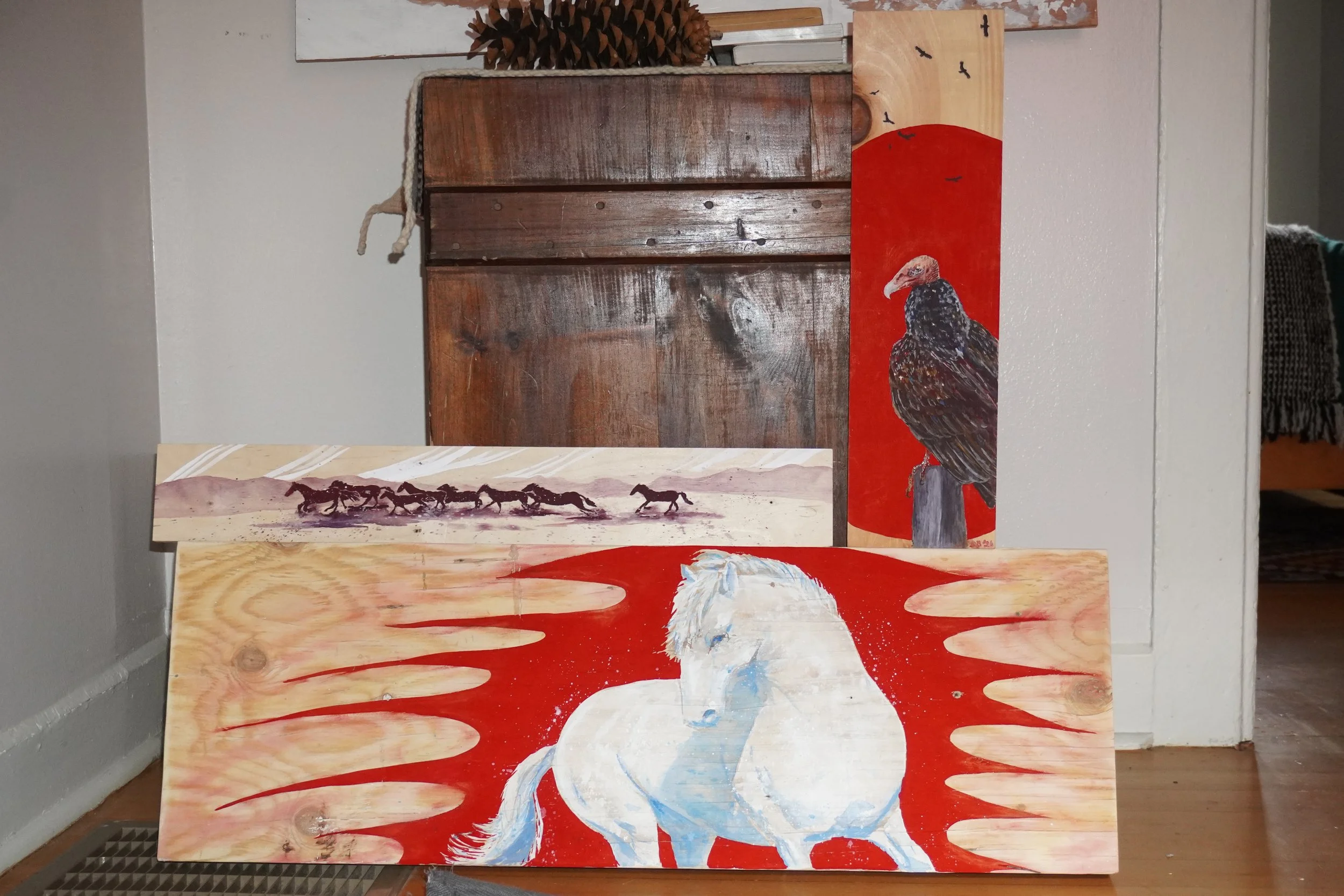 Collection of rustic wildlife paintings featuring a white horse, turkey vulture, running horses, and pinecones leaning against a wooden door