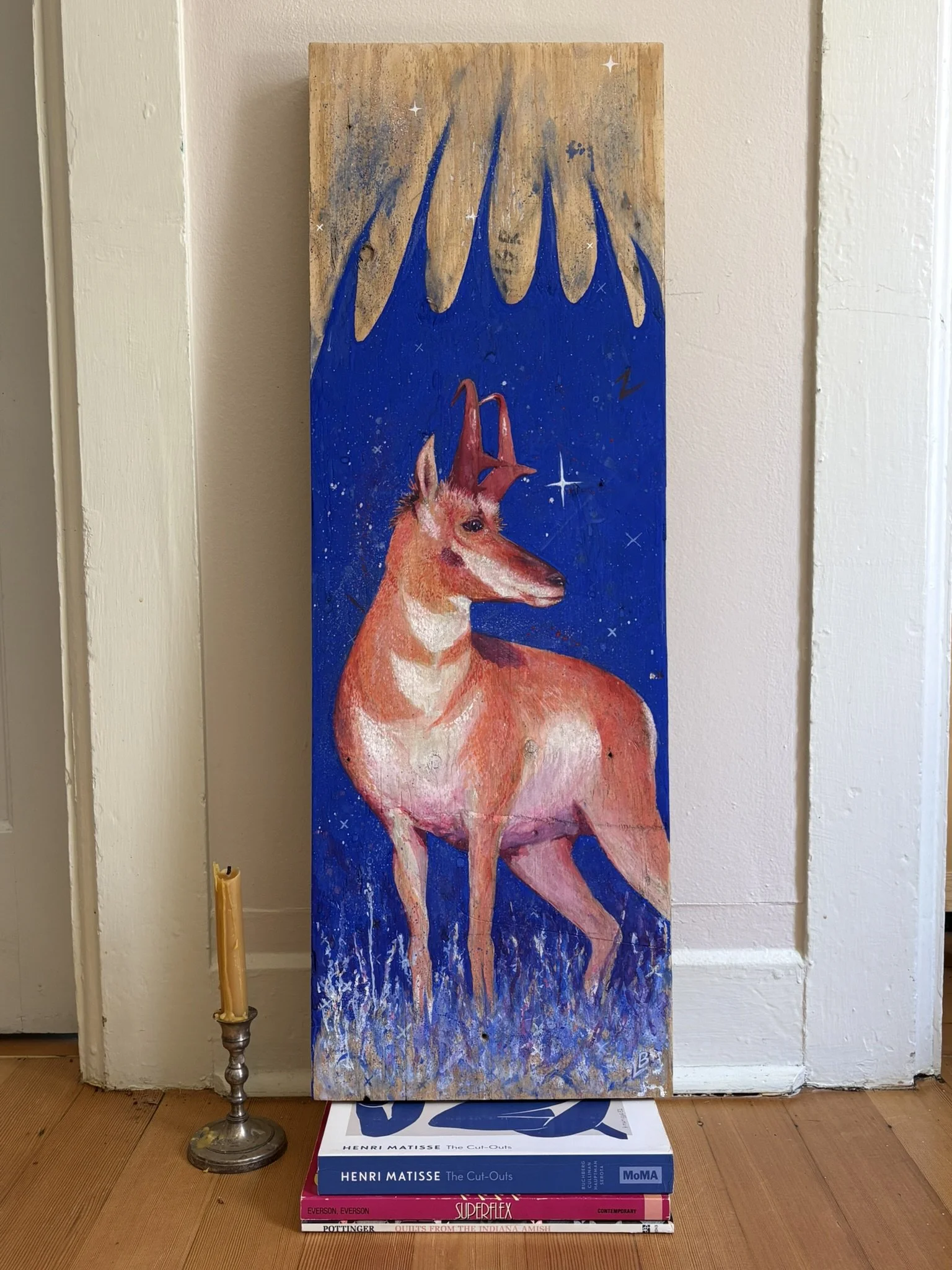 Colorful painting of a Pronghorn Antelope against a starry blue night sky, displayed beside a vintage candlestick and art books.