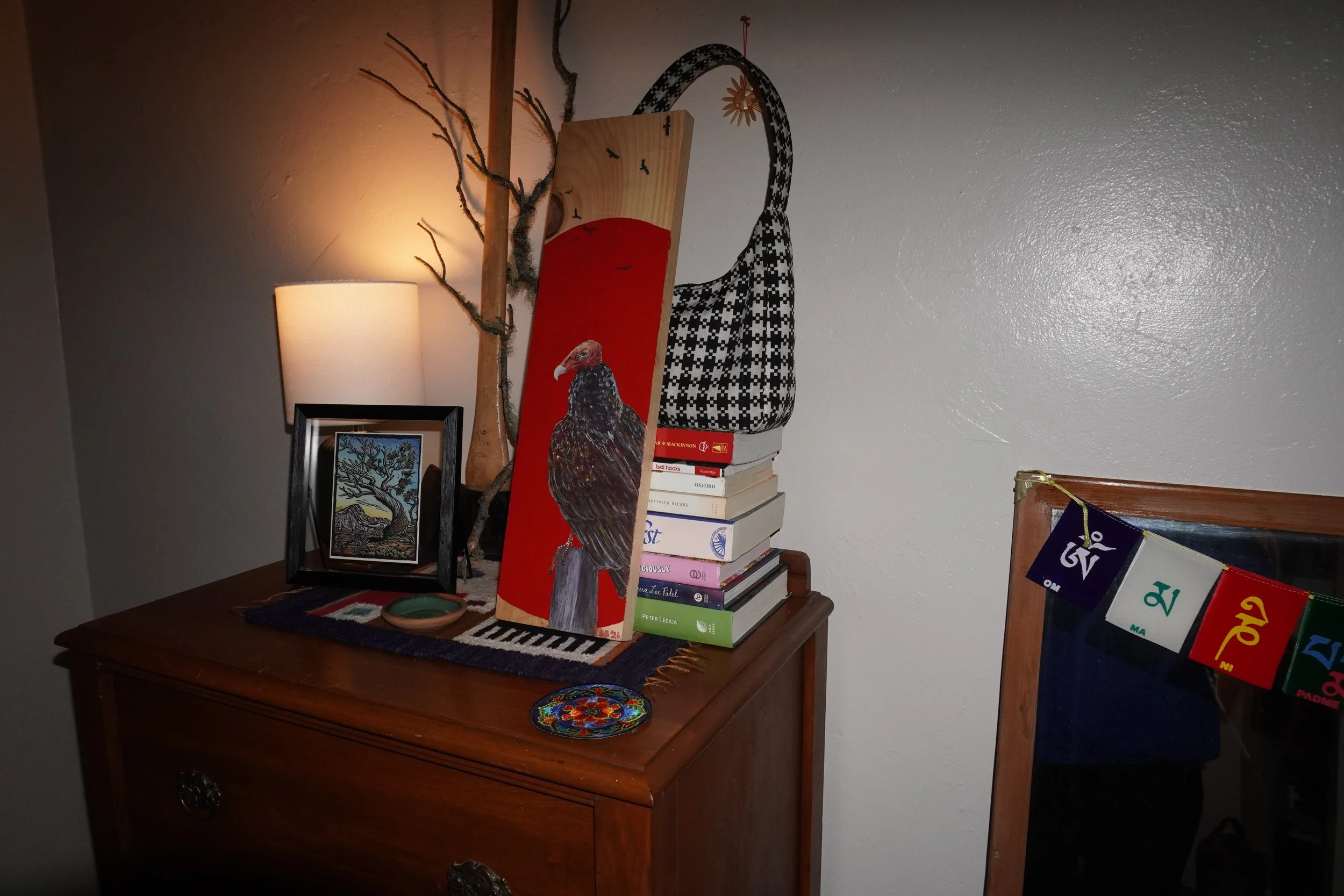 Eclectic wooden dresser display with a lamp, framed artwork, stacked books, turkey-vulture painting, decorative branch, and patterned handbag.
