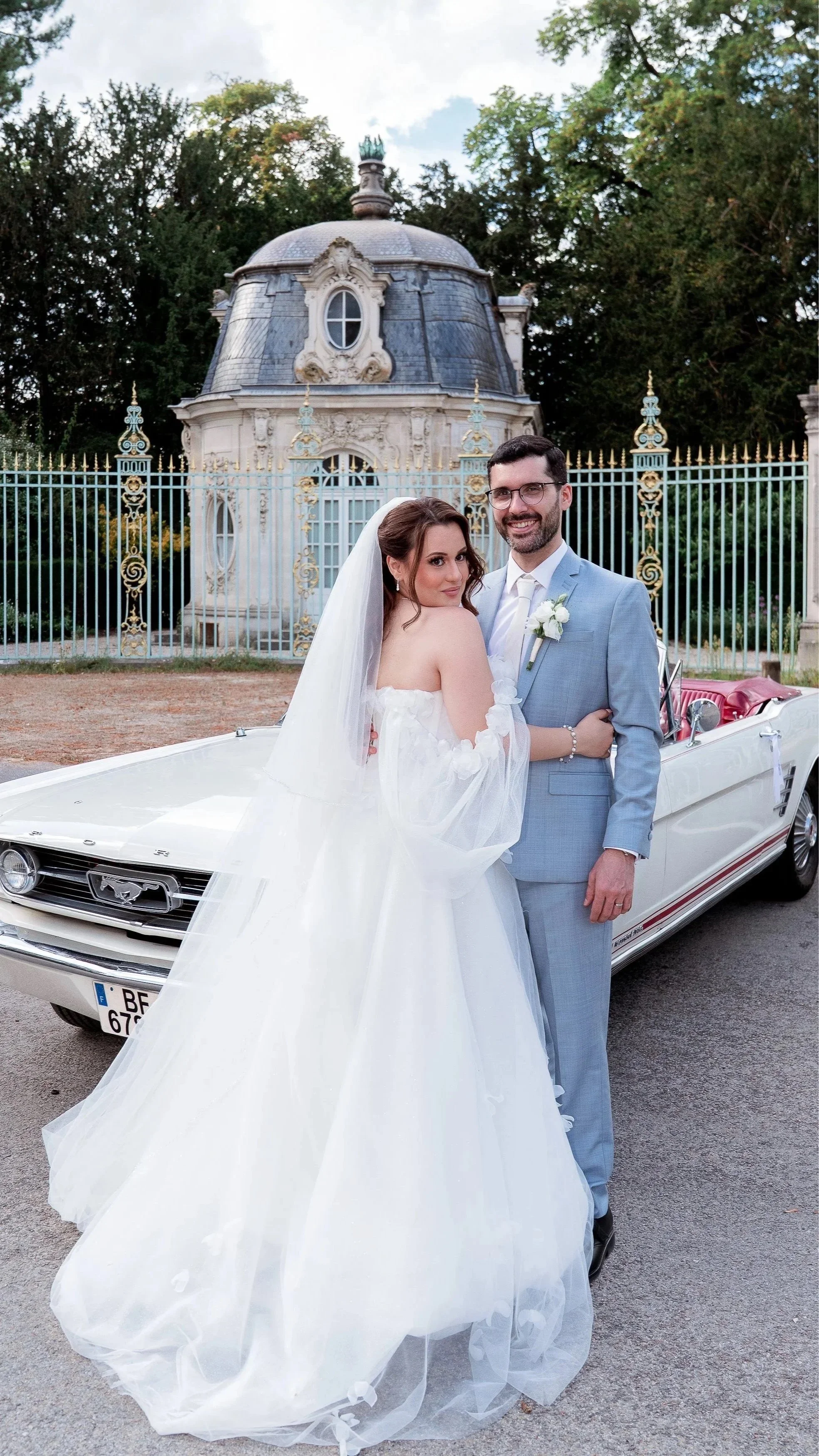 Un couple de mariés pose devant une maison ancienne avec une voiture classique. La mariée porte une robe blanche avec un voile et le marié un costume gris clair avec une boutonnière.