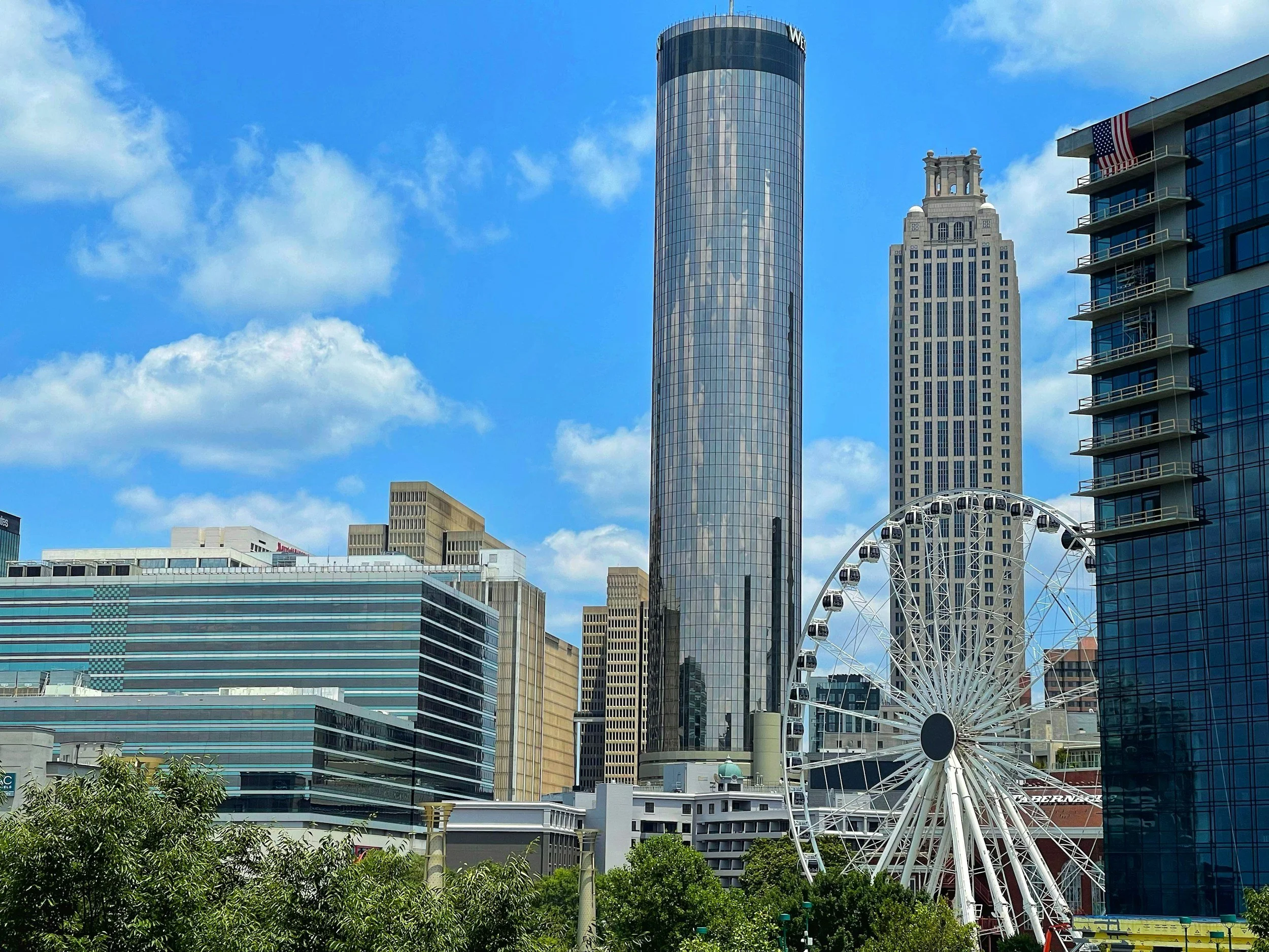 Downtown cityscape with modern skyscrapers, including a tall reflective glass tower, a historic building with a decorative top, a Ferris wheel, and a partly cloudy blue sky.