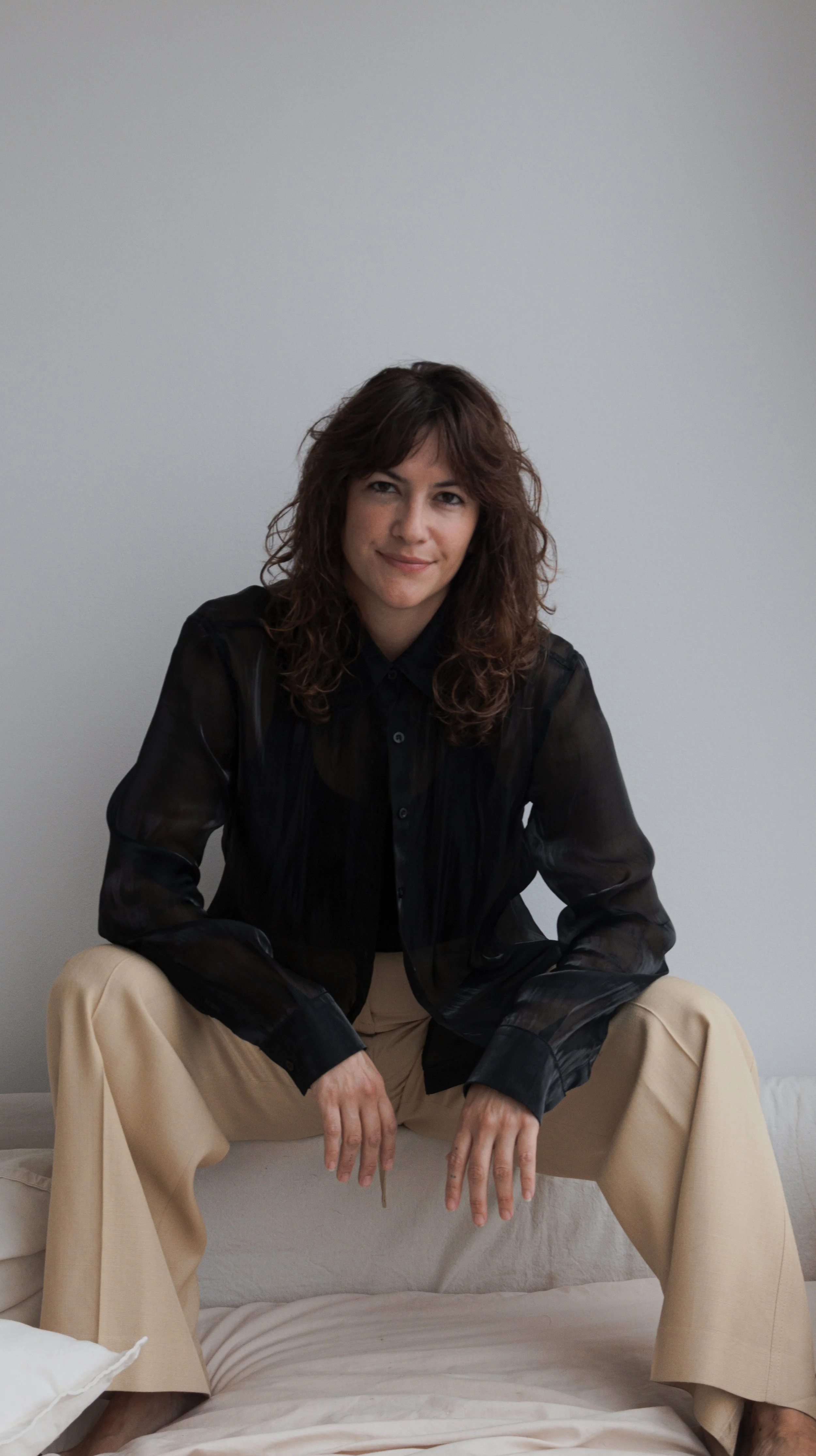 A woman in beige pants and a black shirt sitting on a couch against a plain wall.