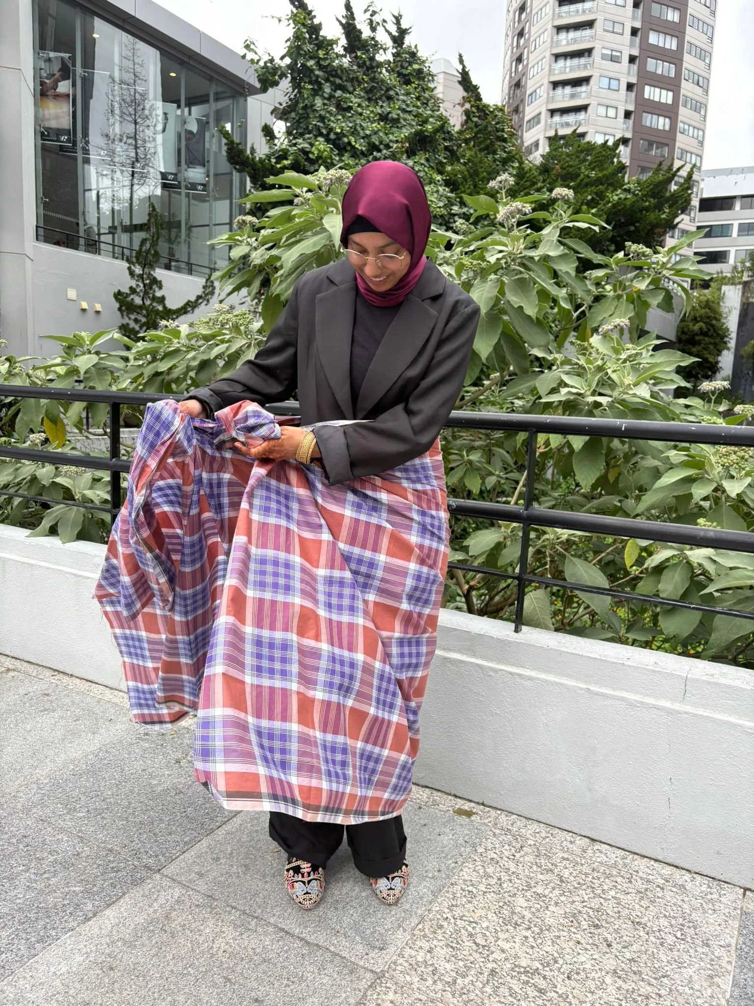 Woman wearing a burgundy hijab, glasses, a black blazer, and a plaid skirt standing outdoors with green foliage and tall buildings in the background.