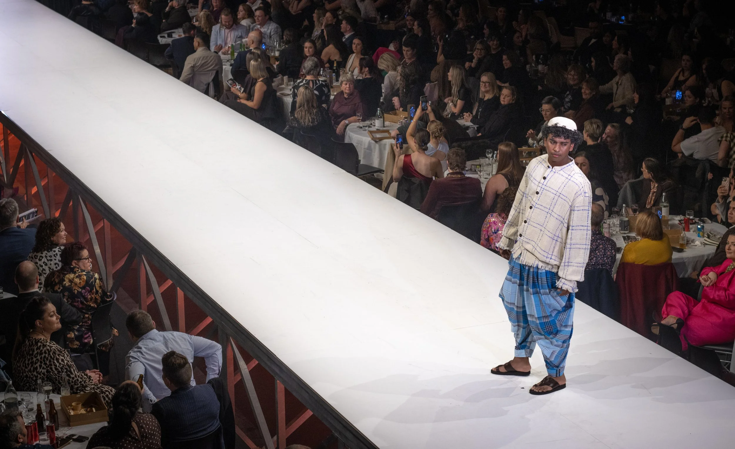 A person in traditional attire walking on a runway during a fashion show, with an audience seated at tables watching and taking photos.