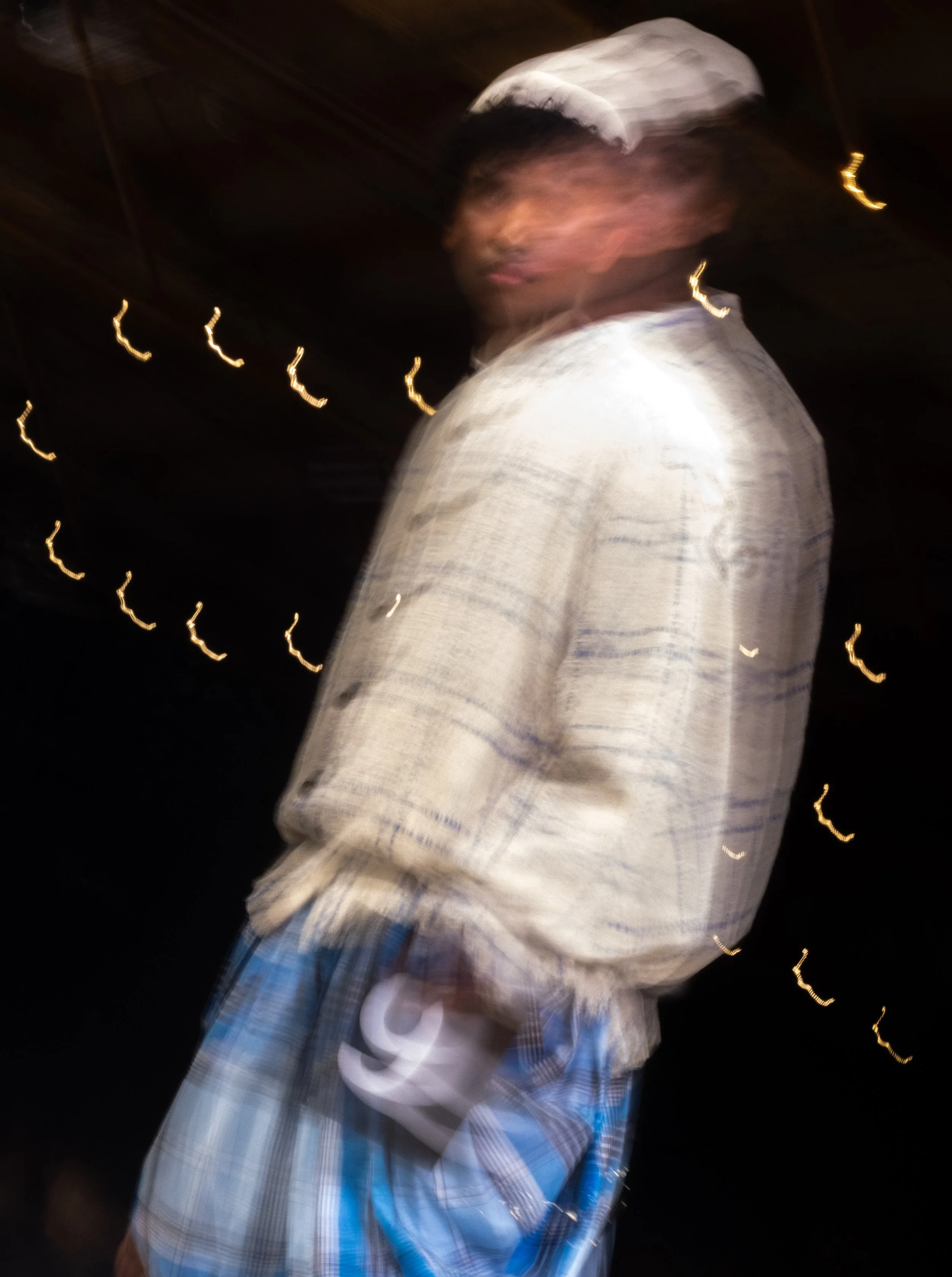 A young man wearing a white cap, a beige shirt, and blue plaid shorts, with motion blur, appears to be in a dark setting with small, bright, curved light streaks in the background.