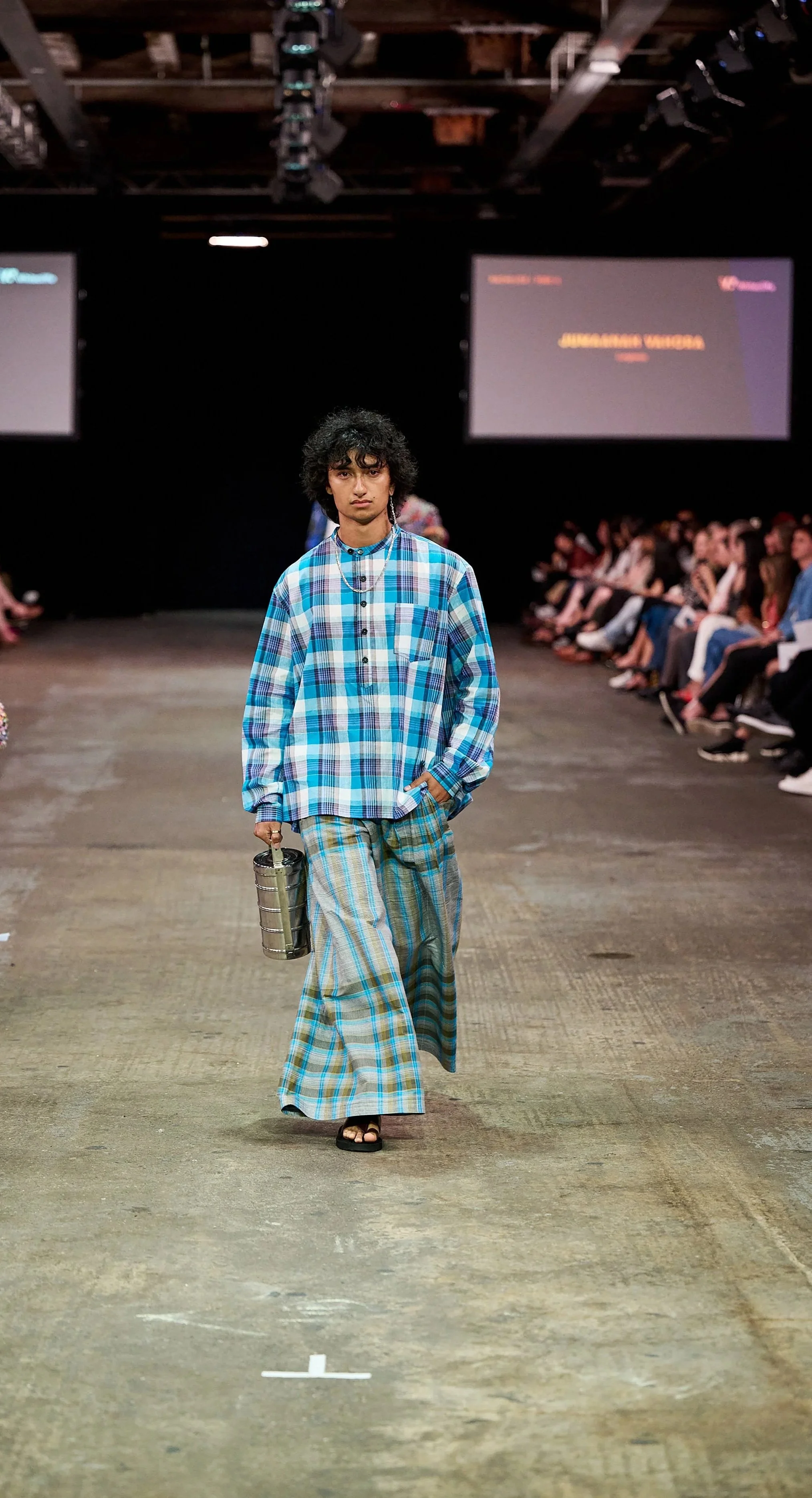 A male model walking down a runway during a fashion show, dressed in a casual plaid shirt, loose plaid pants, and holding a small metallic bag.