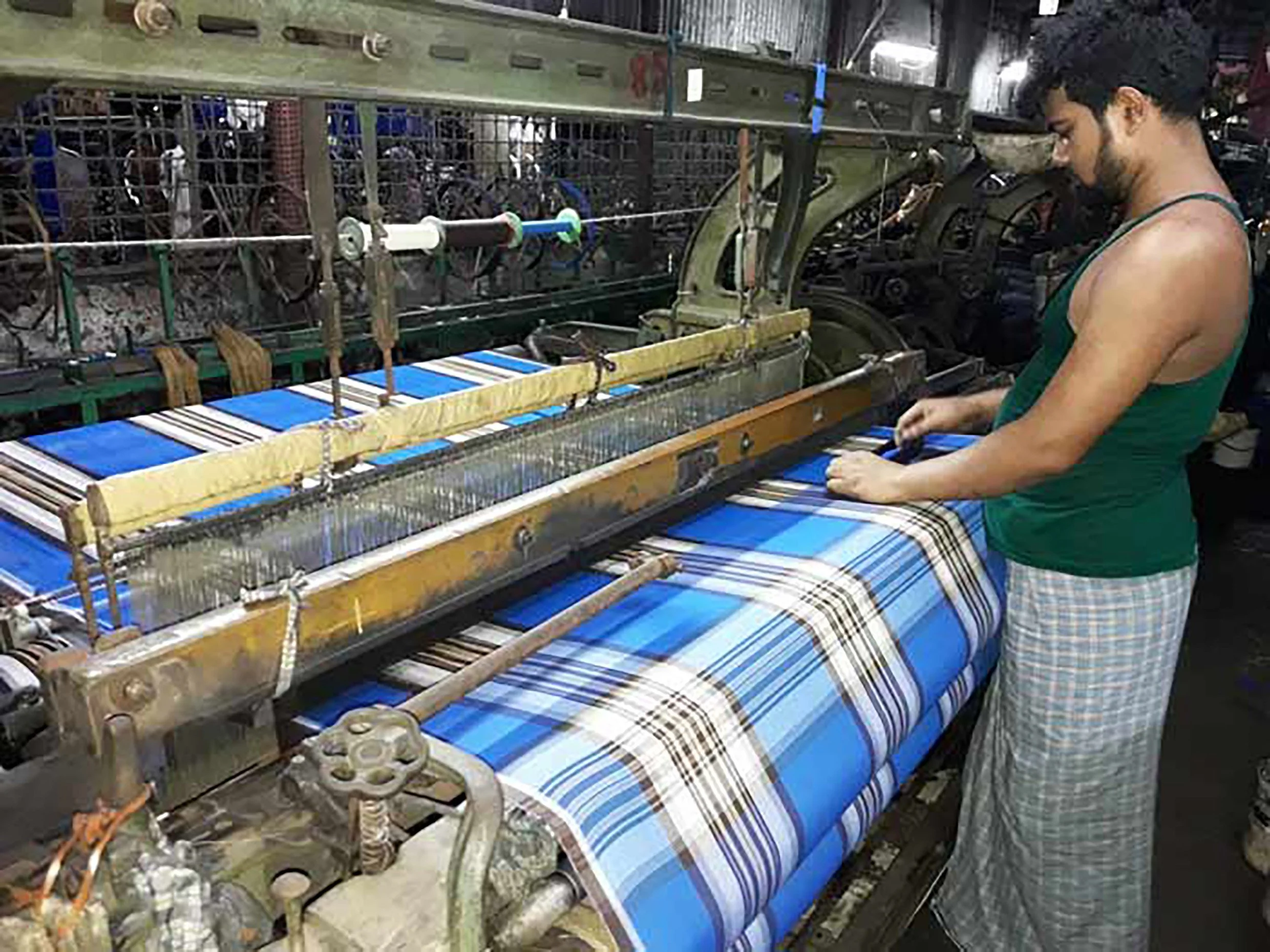 A woman working on a fabric weaving loom with a colorful plaid fabric. She is wearing a green tank top and plaid pants, focused on her task in a workshop surrounded by machinery and woven fabric.