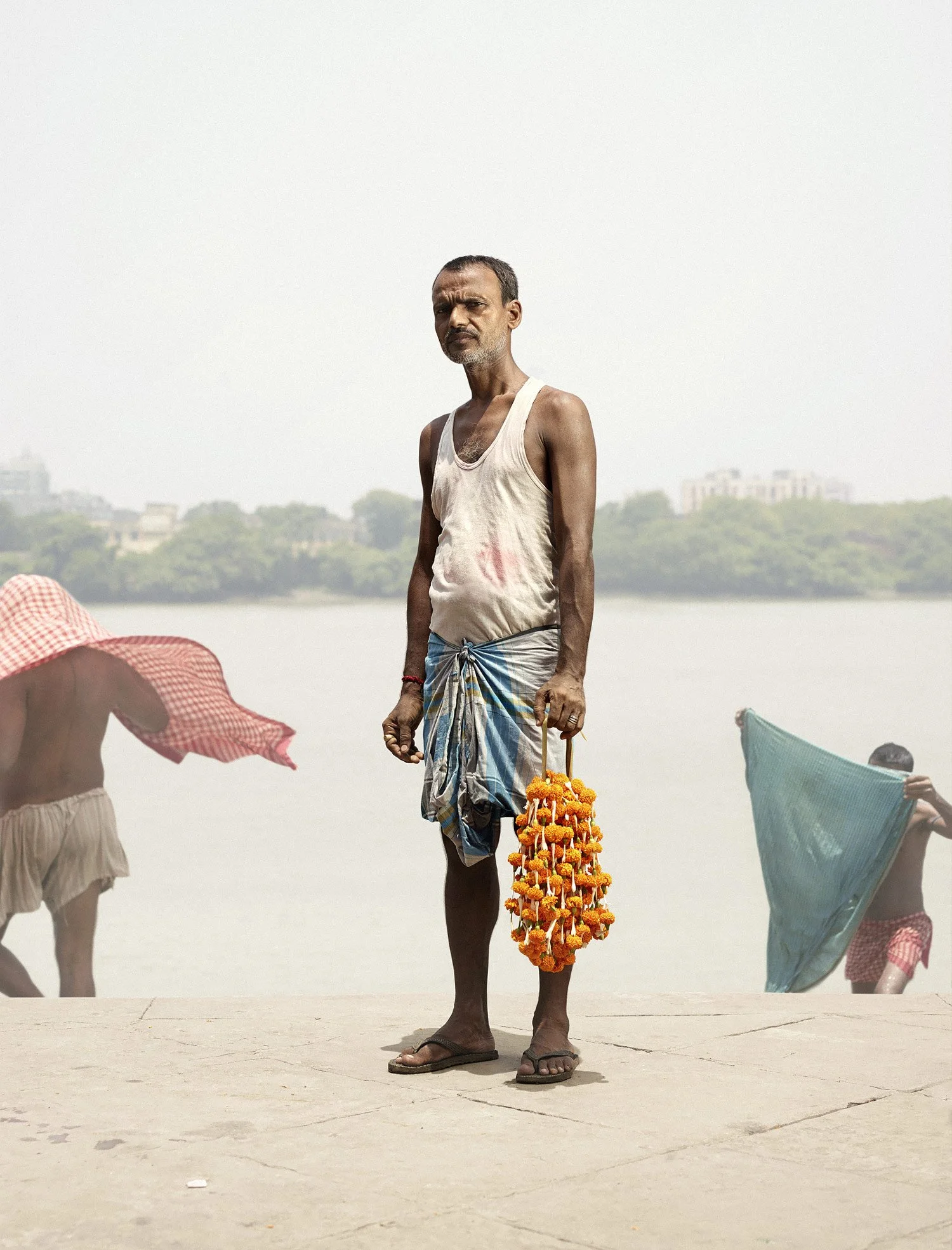 A man with a serious expression standing on a sandy ground, holding a floral garland in one hand, wearing a sleeveless shirt and a traditional cloth around his waist, with two children beside him partially visible, holding umbrellas, in a hazy outdoor setting with a distant view of trees and buildings.