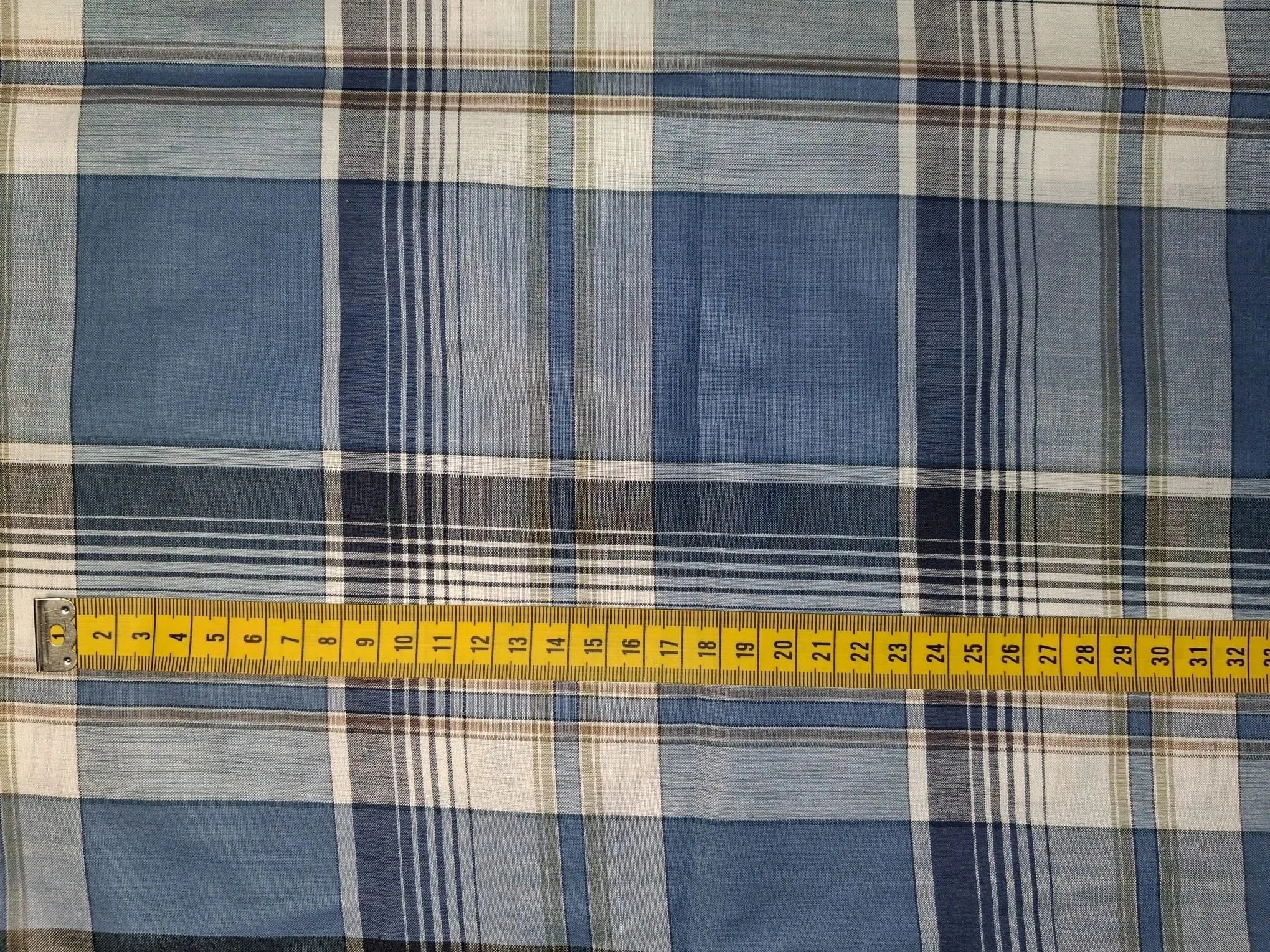 Close-up of a fabric with a plaid pattern in blue, white, beige, and black, with a yellow measuring tape laid horizontally across the bottom edge.