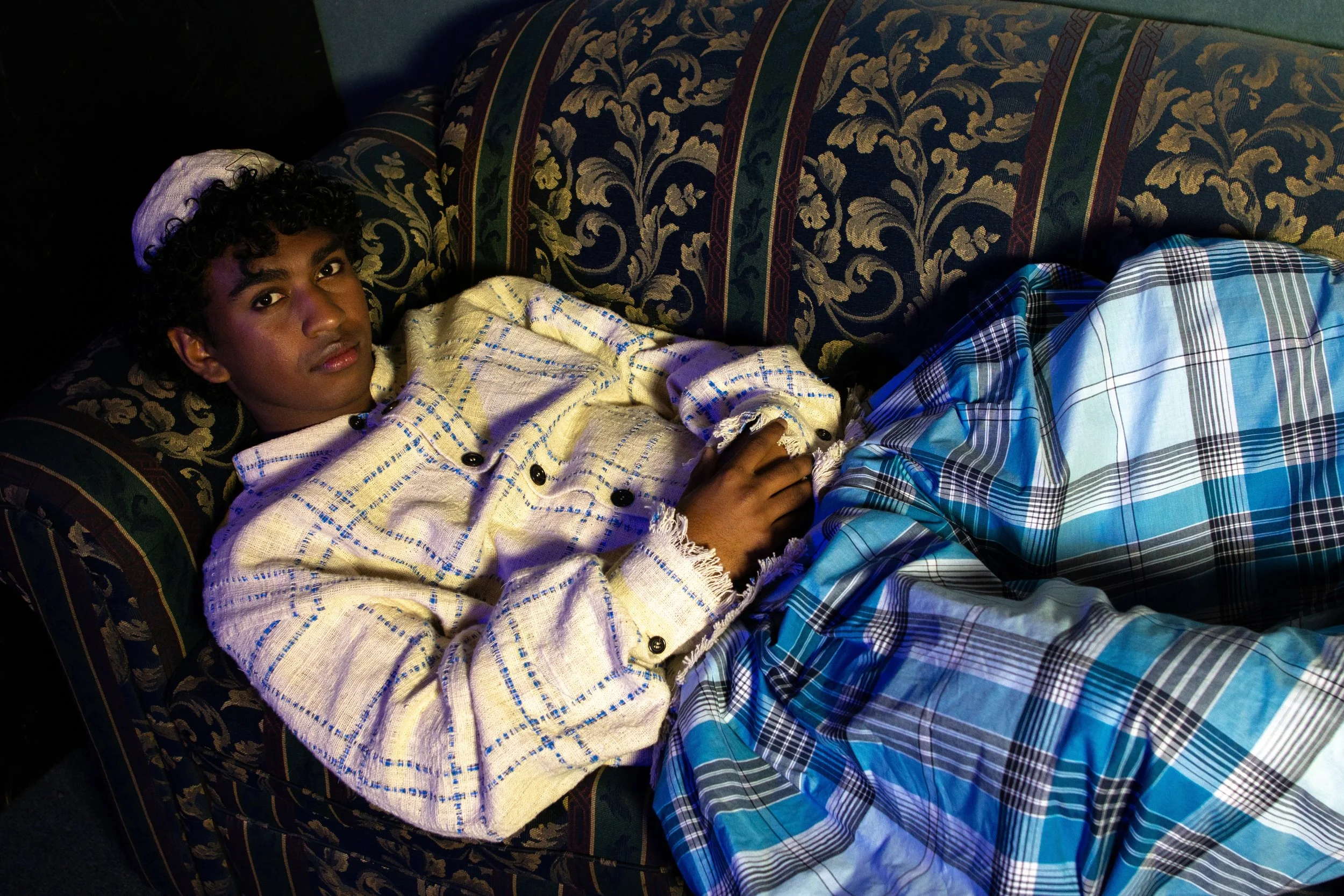 Young man with curly hair lying on a sofa, wearing a light-colored jacket and plaid pants, looking at the camera.