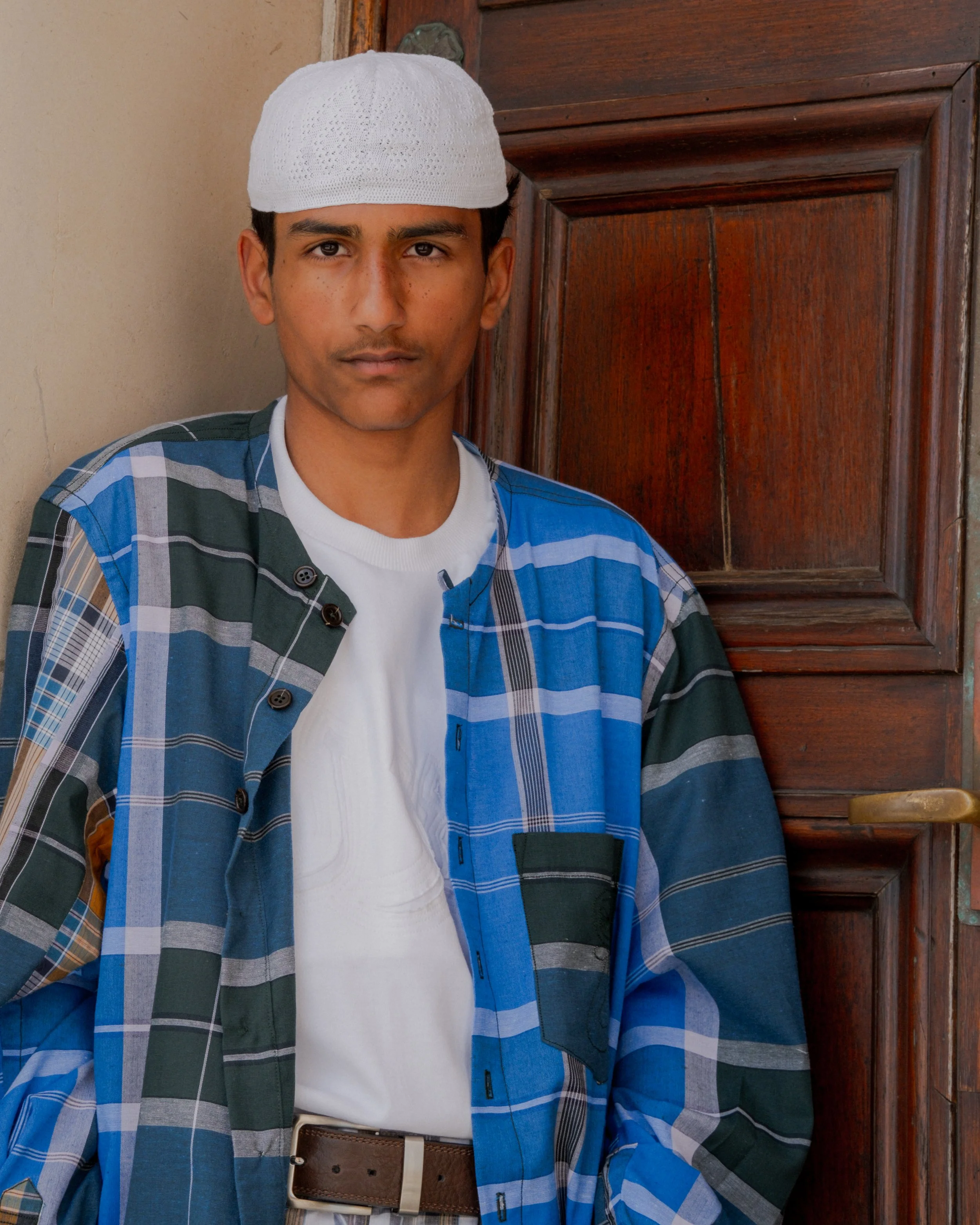 A young man with tan skin and dark hair, wearing a white prayer cap, a white T-shirt, a plaid shirt in shades of blue, gray, and cream, and a brown belt with beige pants, standing next to a wooden cabinet with a drawer on the right side.