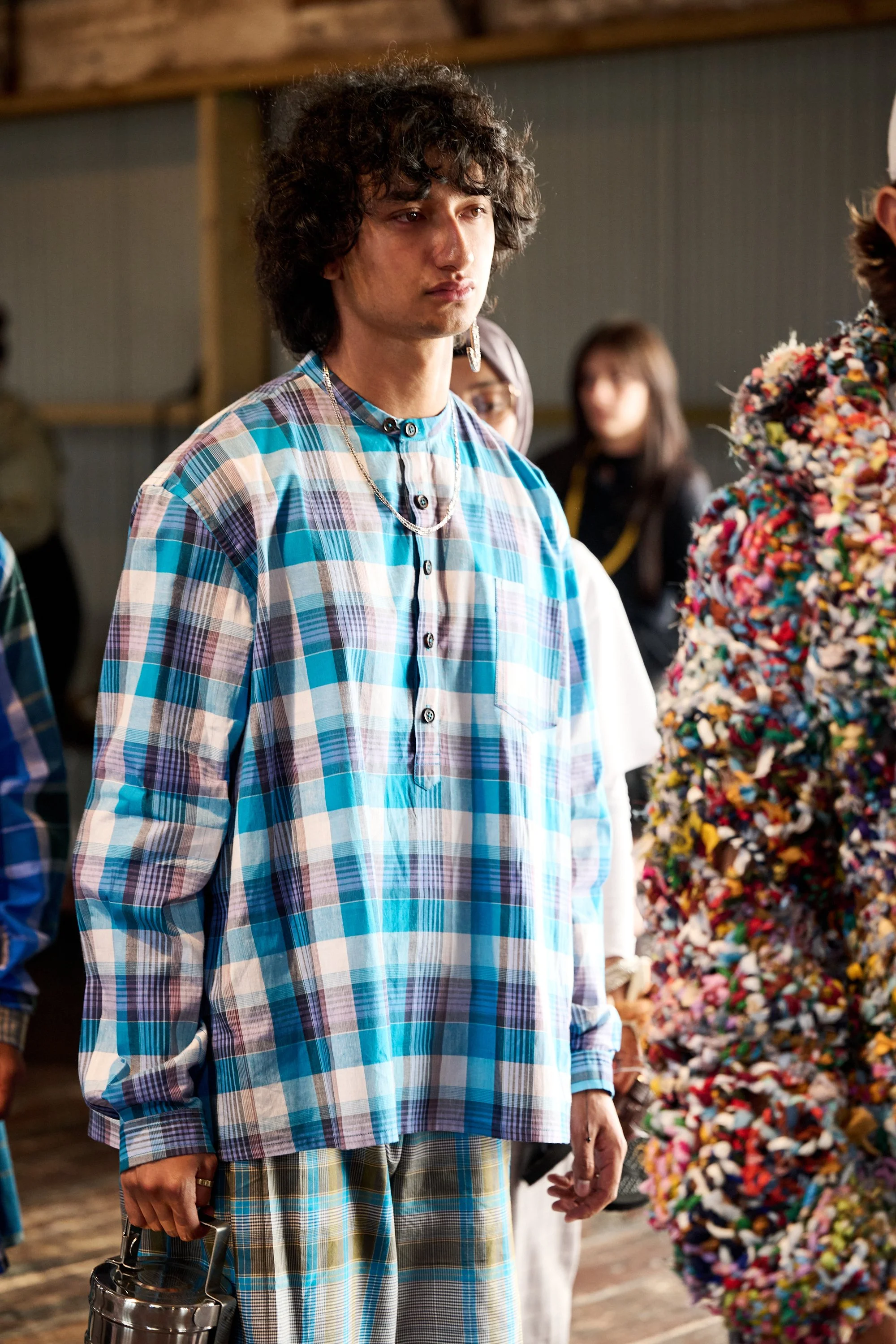 A young man with curly dark hair wearing a blue and white plaid shirt, holding a clear bag, standing in an indoor setting with other people and a colorful, textured backdrop.
