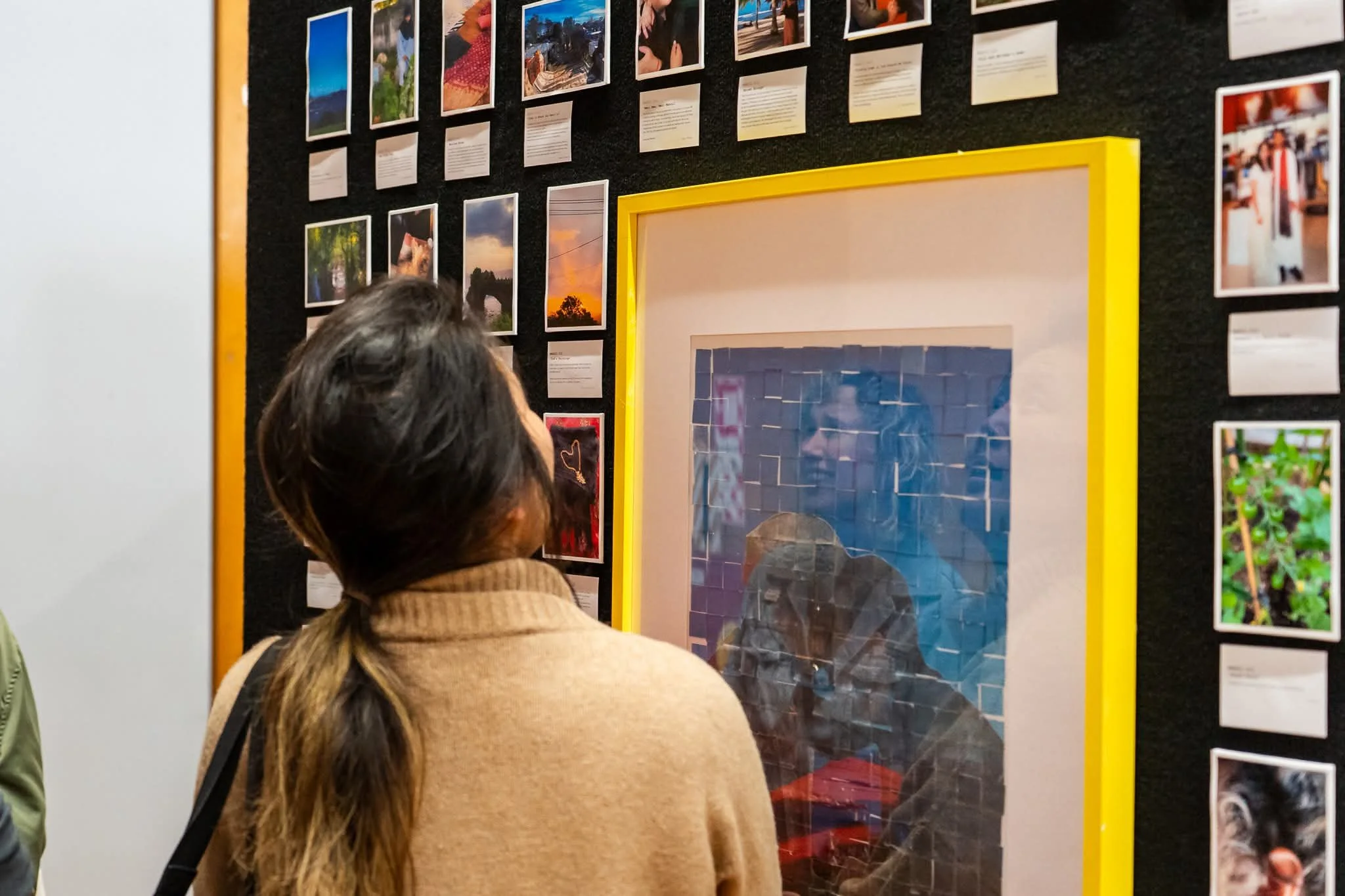 A person with long hair in a ponytail wearing a beige sweater is looking at a photo display on a black wall, which features various landscape and outdoor photographs and text descriptions. Part of another person's arm is visible on the left side of the image.