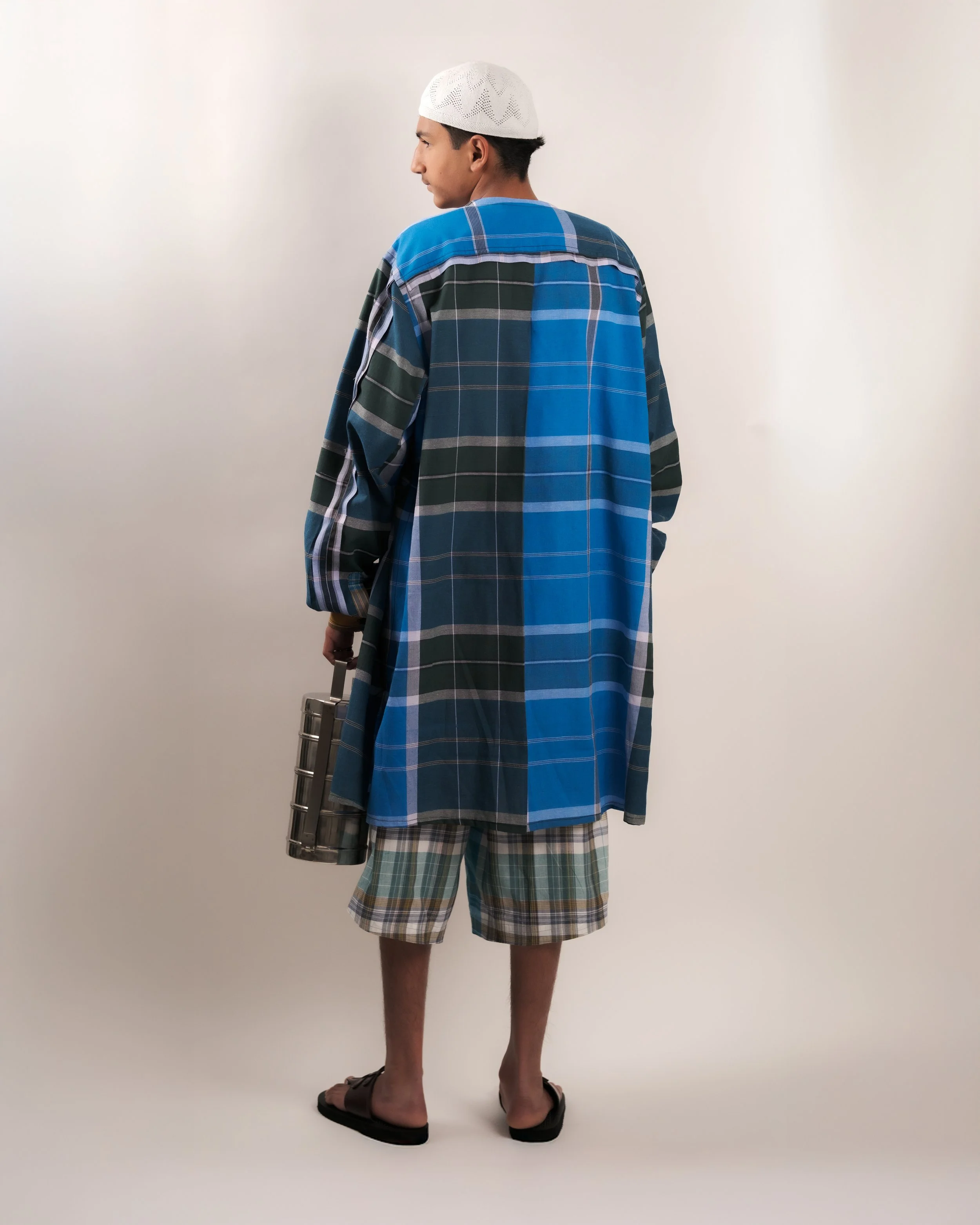 A man dressed in a long, multi-colored plaid shirt, paired with plaid shorts, wearing a white knitted cap and brown sandals, standing against a plain white wall.