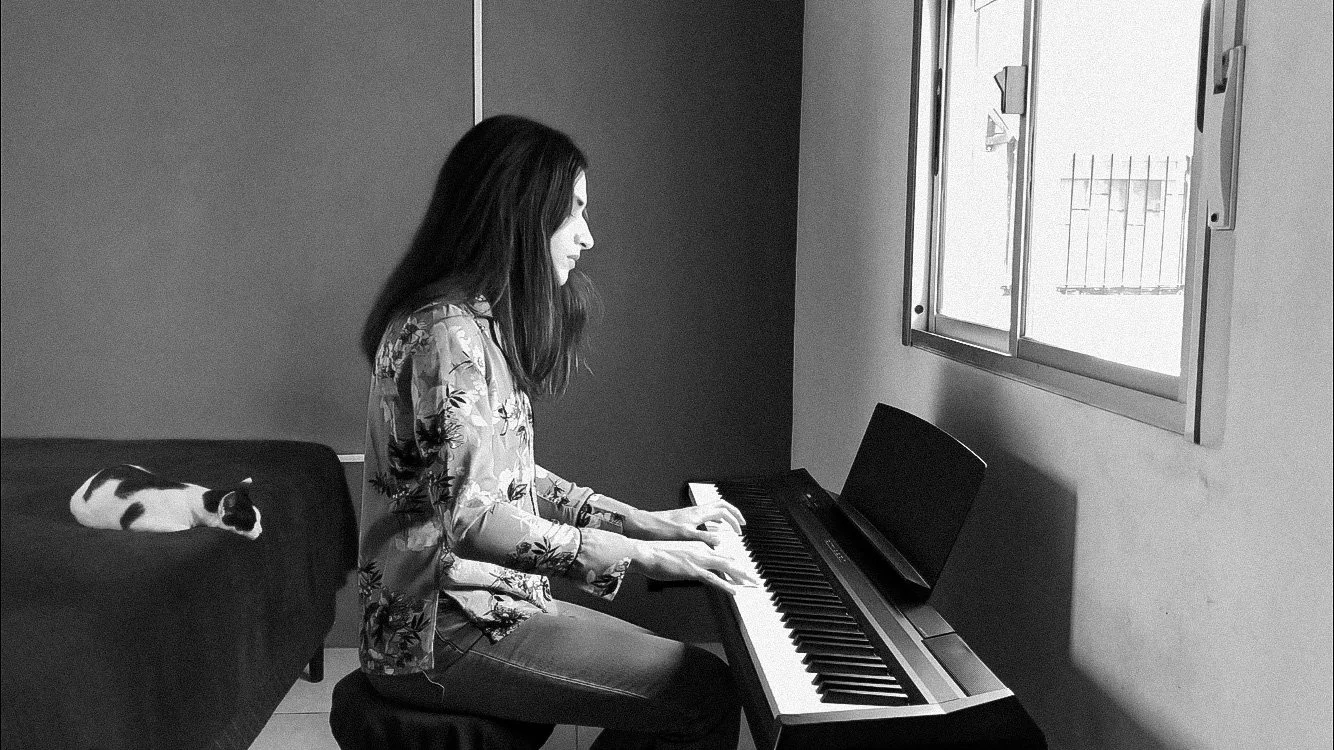 Chico con cabello largo tocando el piano mientras un gato de pelaje blanco y negro duerme al fondo