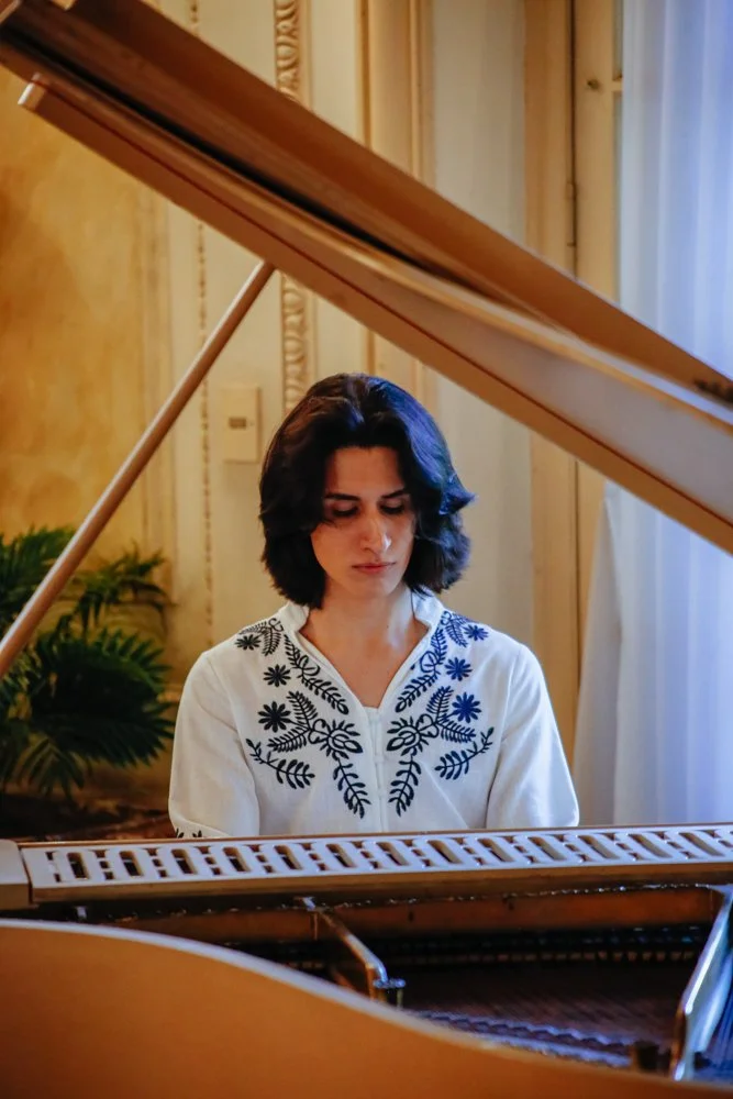 Chico tocando un piano en una habitación con cortinas blancas y plantas verdes.