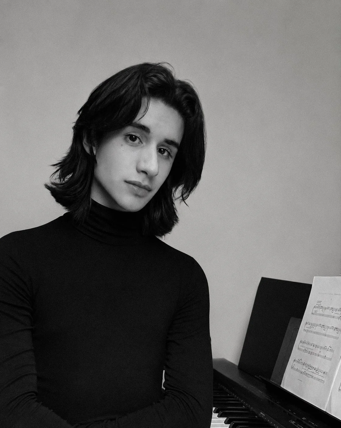 Chico con cabello oscuro y liso, vestido con suéter negro, sentado frente a un piano con partitura, en un entorno gris y sencillo.