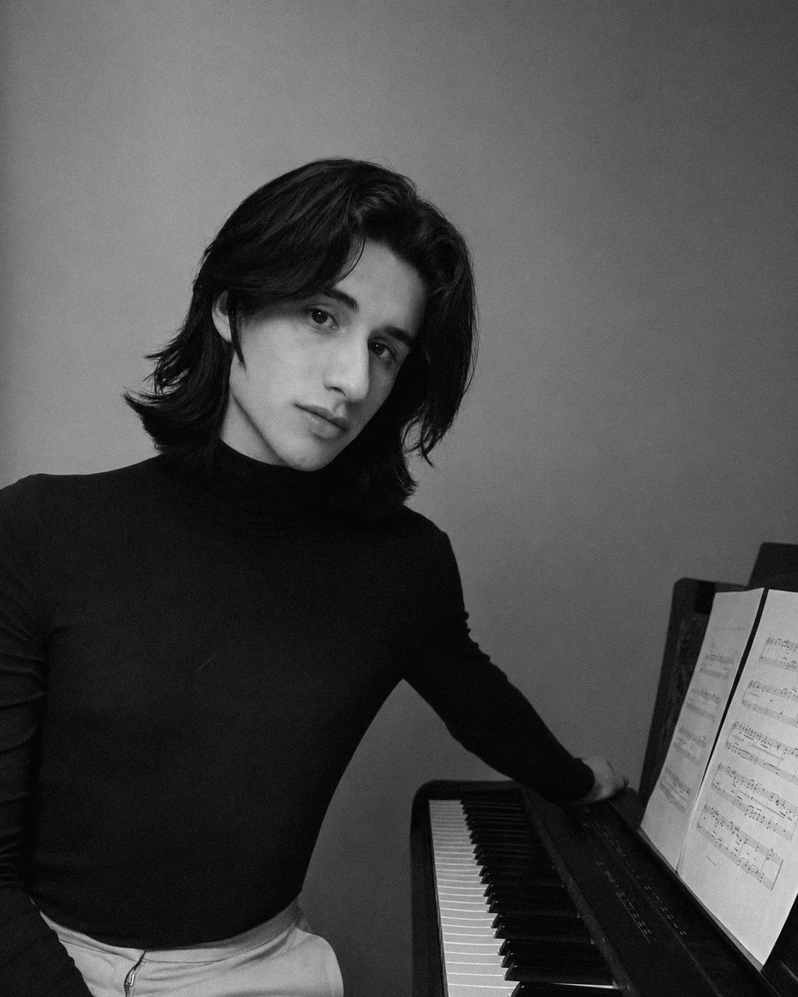 Chico con cabello largo y oscuro tocando un piano, en un fondo gris, en una fotografía en blanco y negro.