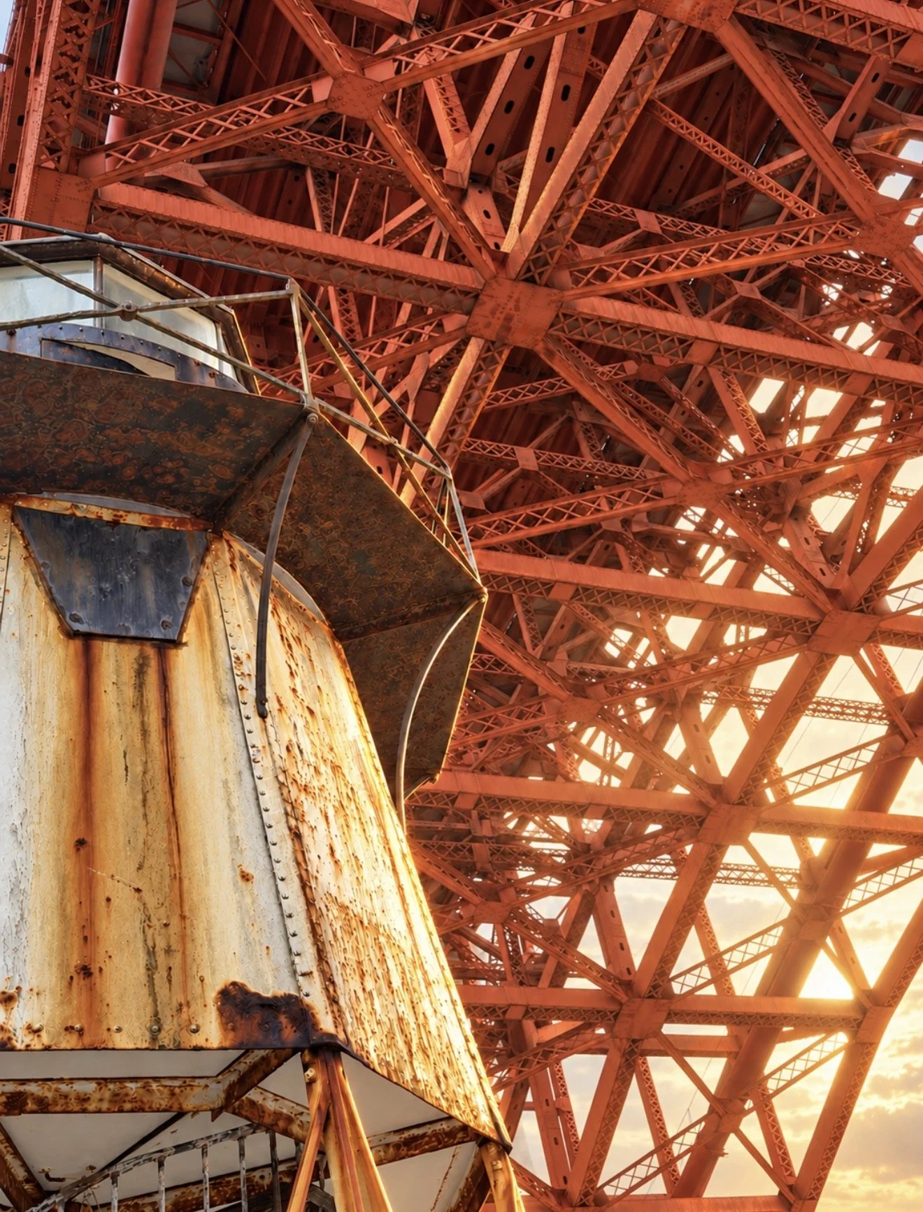 Close-up view of a rusted, weathered carnival ride or Ferris wheel with a background of a large red metal structure, likely part of the same amusement park ride, during sunset.