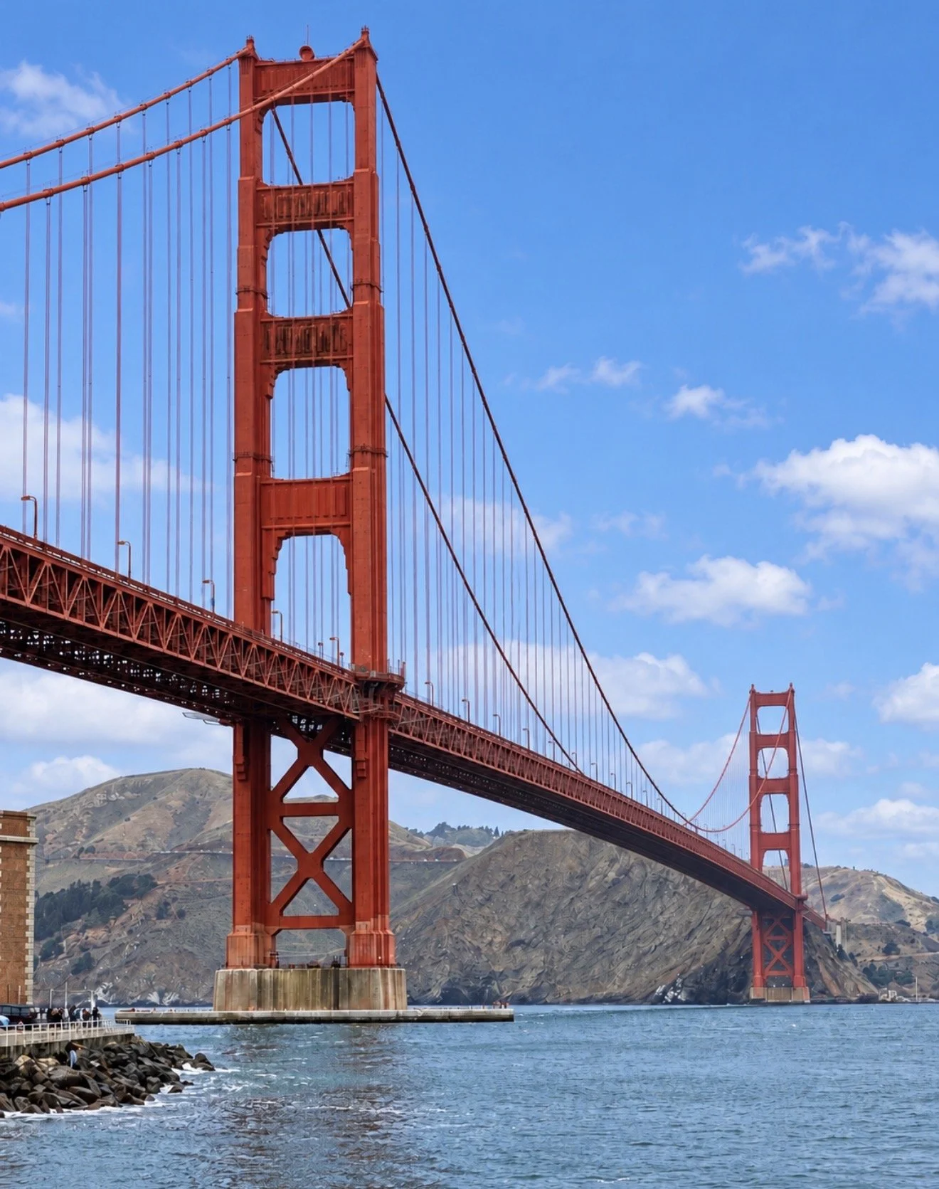 A view of the Golden Gate Bridge in San Francisco, California during the day, with blue sky and clouds in the background. The bridge's towers are red-orange and the water below is blue with a rocky shoreline in the foreground.