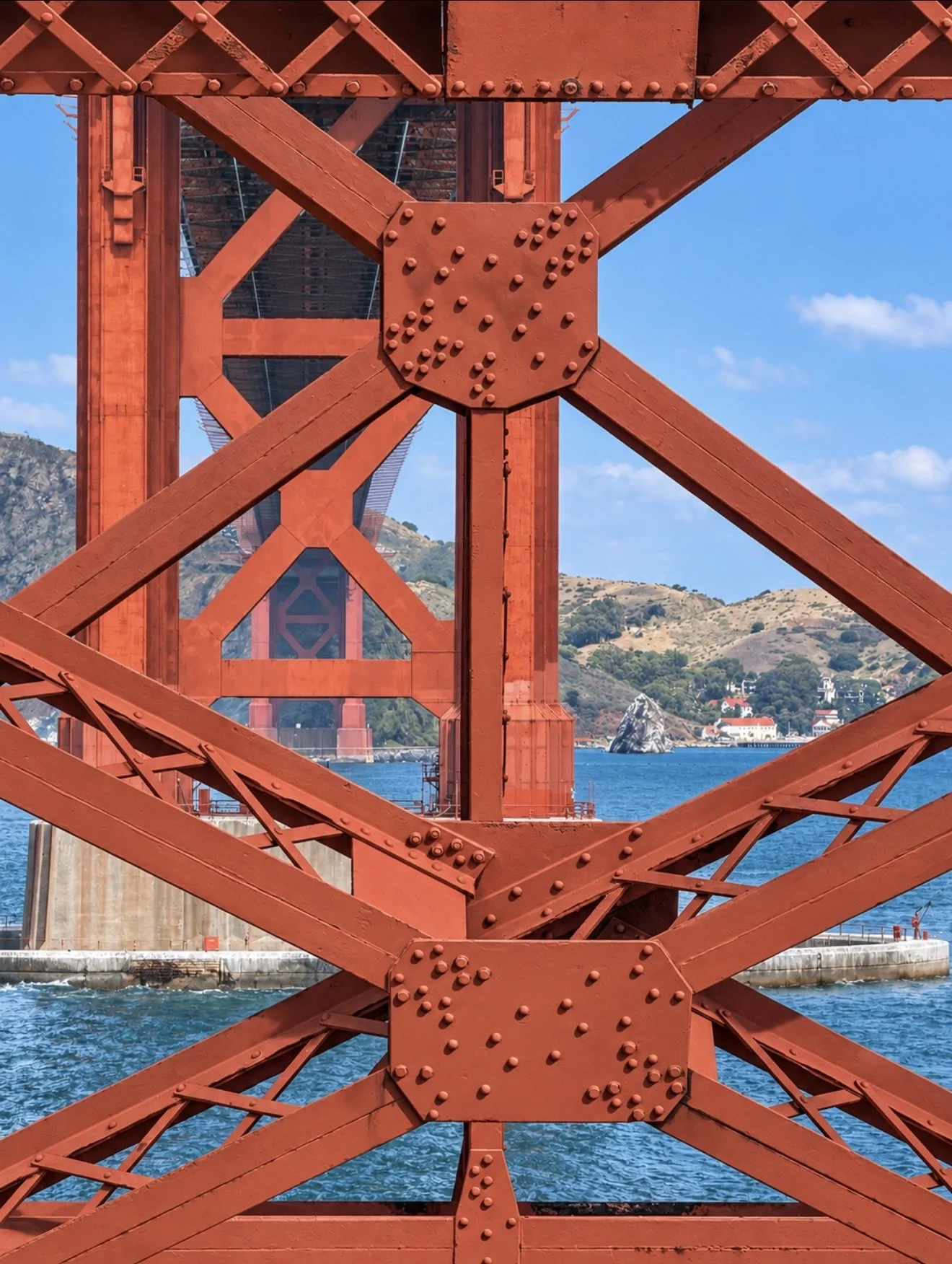 Close-up view of the red steel structure of the Golden Gate Bridge with a landscape and water visible in the background.