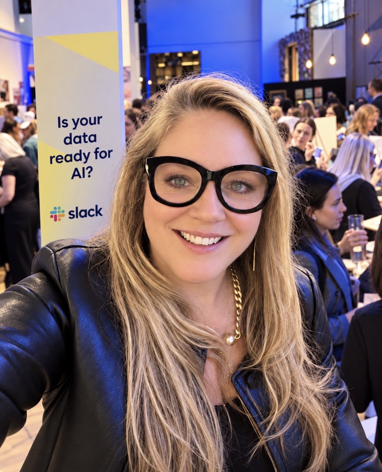 A woman with long blonde hair, wearing glasses, a black leather jacket, and jewelry, taking a selfie at a crowded event. In the background, a banner asks, 'Is your data ready for AI?' with Slack's logo.