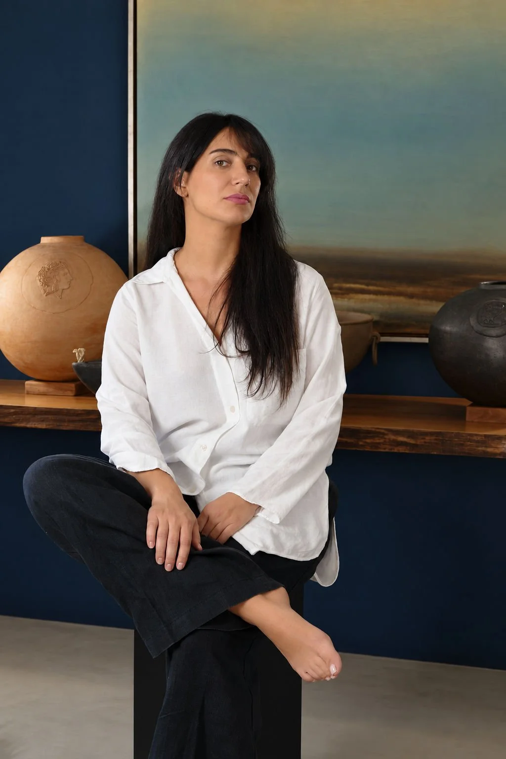 A woman with long dark hair wearing a white shirt and dark pants sitting with one leg crossed over the other in front of a dark blue wall with decorative pottery and a landscape painting.