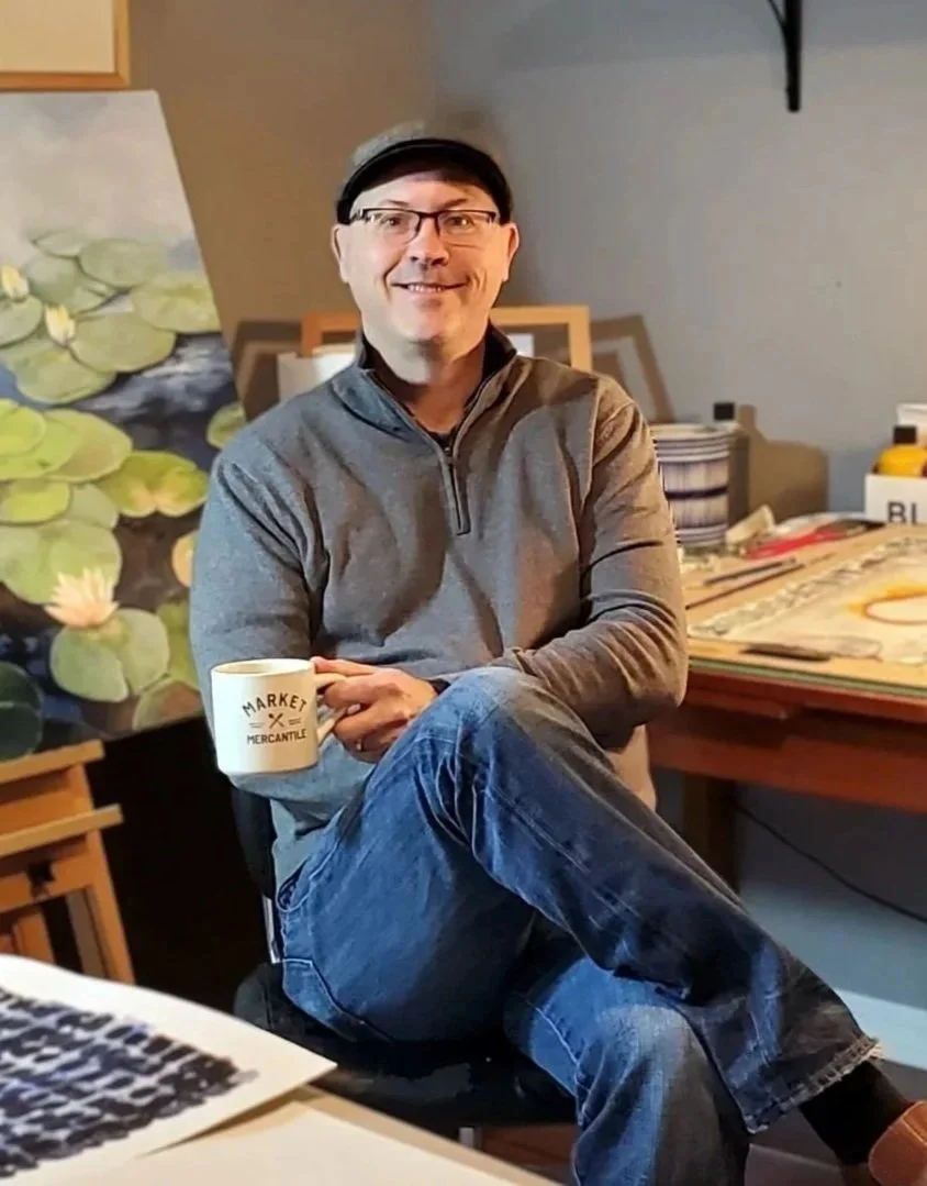 Photo of artist, Bruce Black in his art studio.  He sits with coffee in hand and a painting of water lilies behind him.