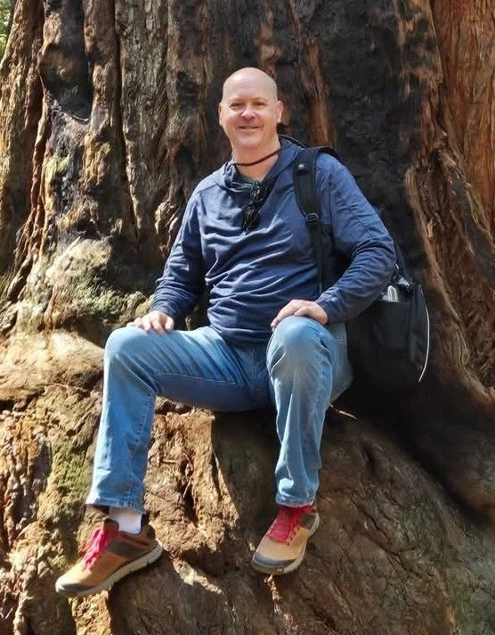 Bruce Black sitting on section of a redwood tree in Northern California near the coast