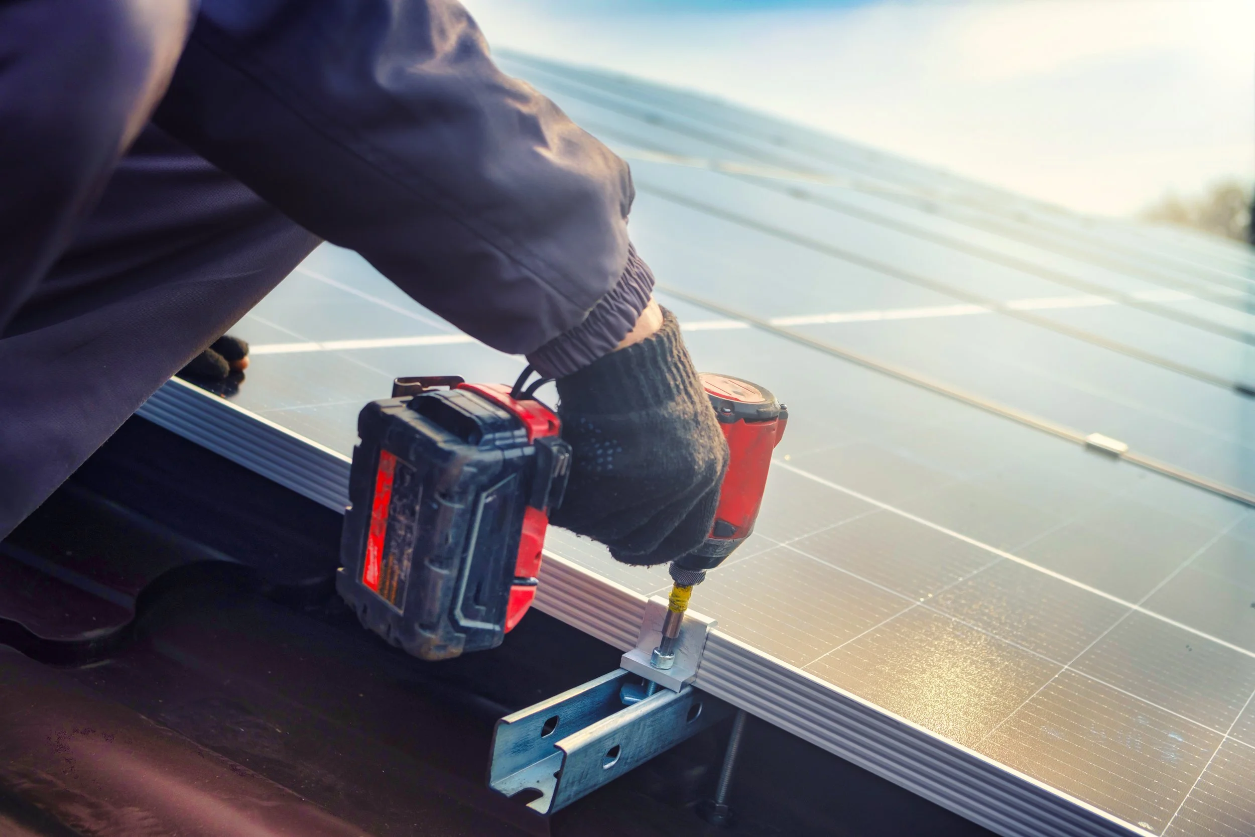 Close-up of a worker installing solar panels, using a power drill on a solar panel array outdoors during daytime.