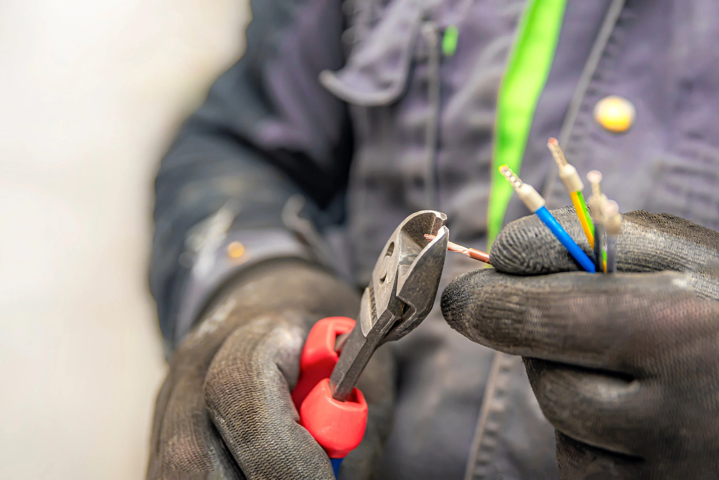 A worker wearing gloves is using pliers to hold electrical wires with colored insulation, likely as part of an electrical repair or assembly.