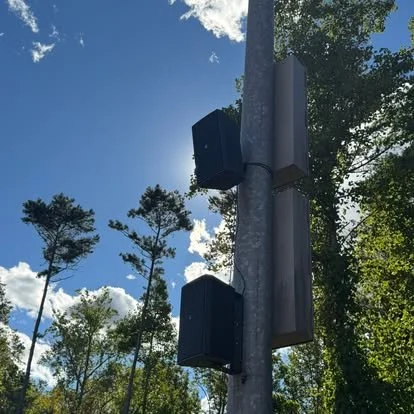 A pole with three black rectangular speaker or sensor boxes in a wooded outdoor setting with trees and a blue sky with clouds.