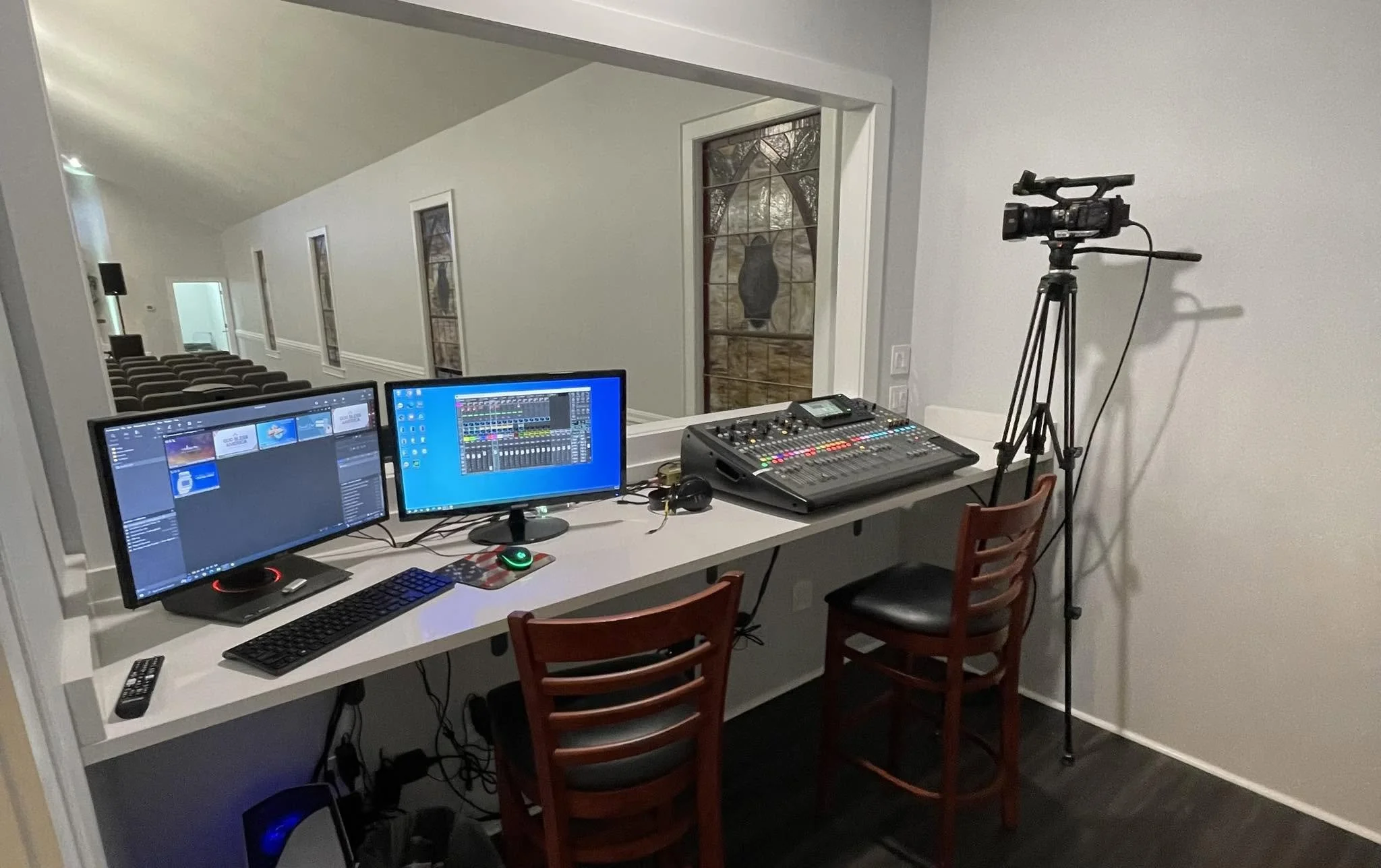A home recording or editing space with two computer monitors, a keyboard, a mouse, a remote, a sound mixer, a camera on a tripod, and chairs in front of the desk. There are multiple windows behind the workspace.
