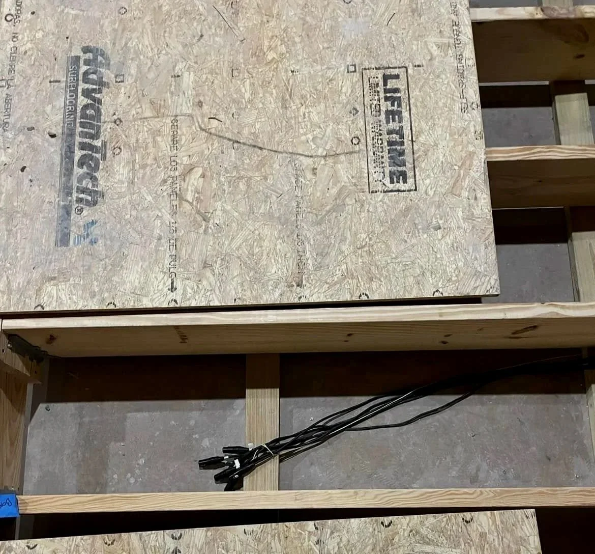 A partially assembled piece of furniture or structure with plywood panels and a wooden frame, featuring some electrical cords on the floor beneath.