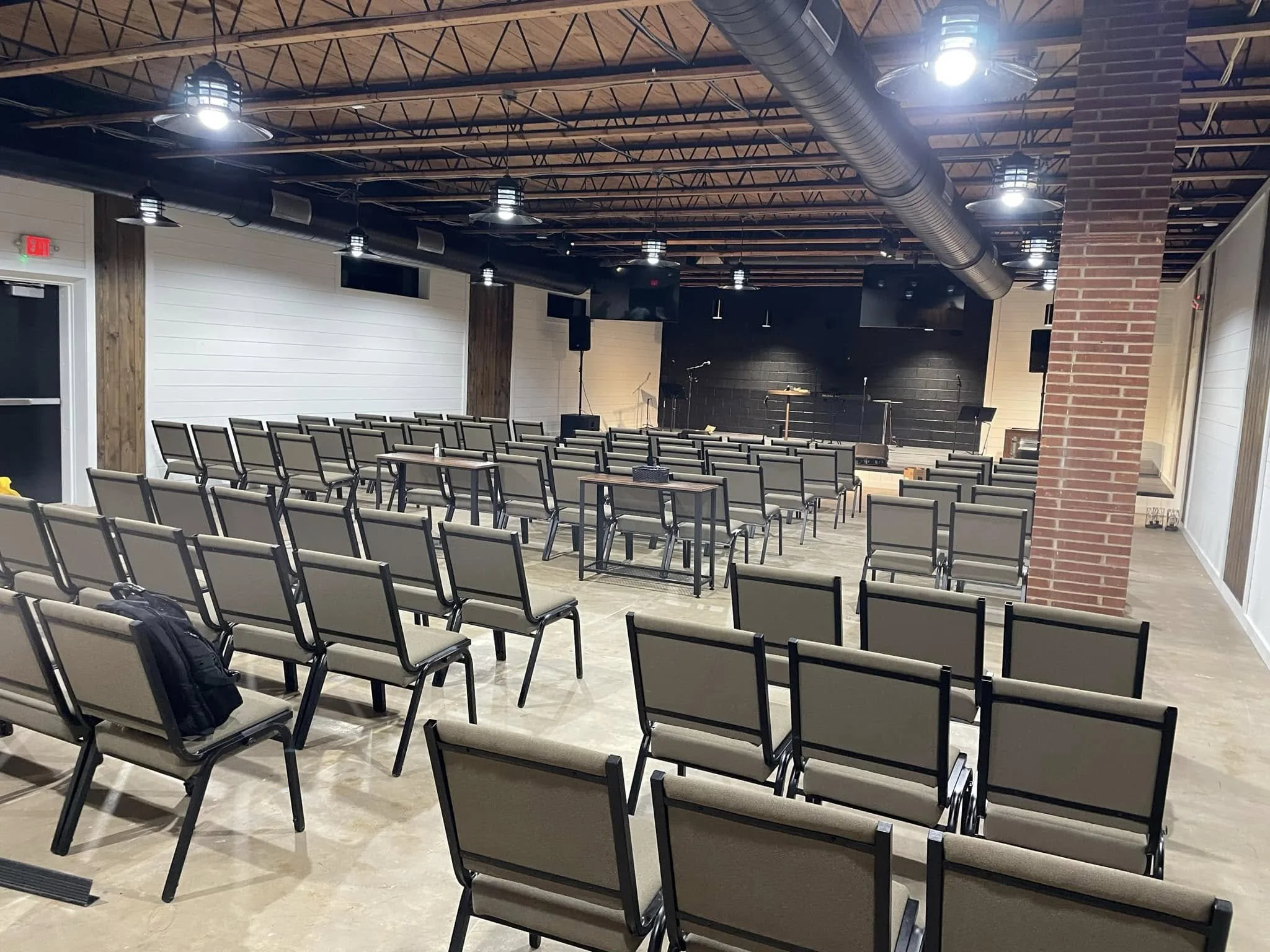 Empty seated chairs facing a stage with microphones and music stands in a spacious auditorium with a black back wall and exposed wooden ceiling.