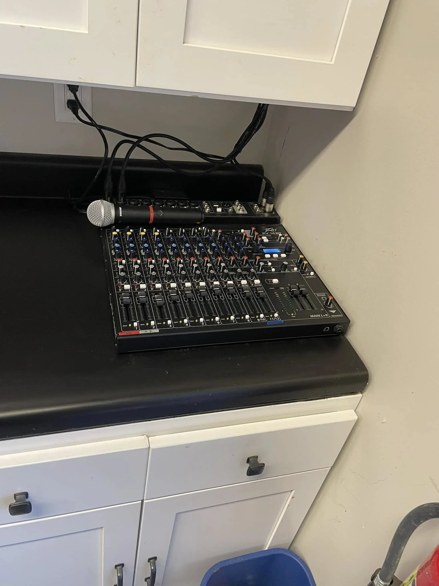 A microphone resting on an audio mixer placed on a white countertop, with cords connected to the mixer. The setup is near a white cabinet and wall.