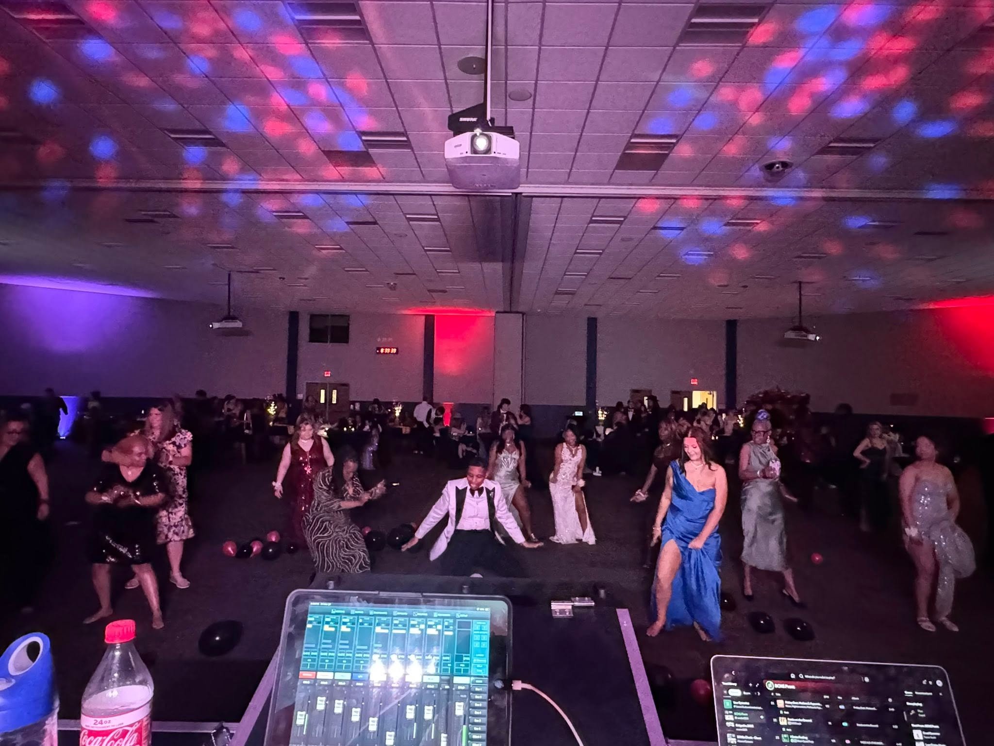 Dance party with people dancing on a dark floor in front of a DJ booth with lighting and colored spotlights on the ceiling.