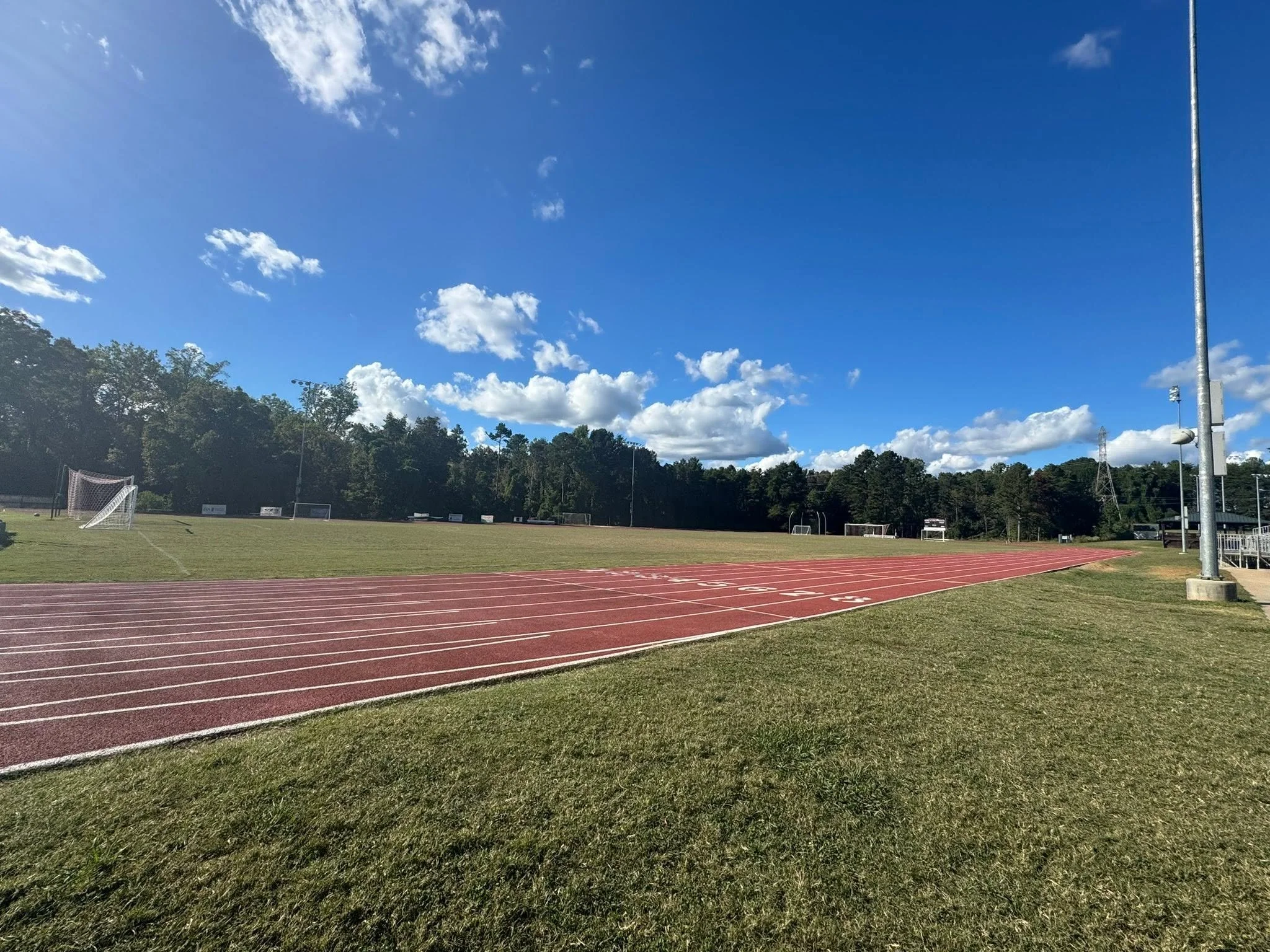 An outdoor athletic track with red lanes, soccer goals, and a field, surrounded by trees under a blue sky with clouds.
