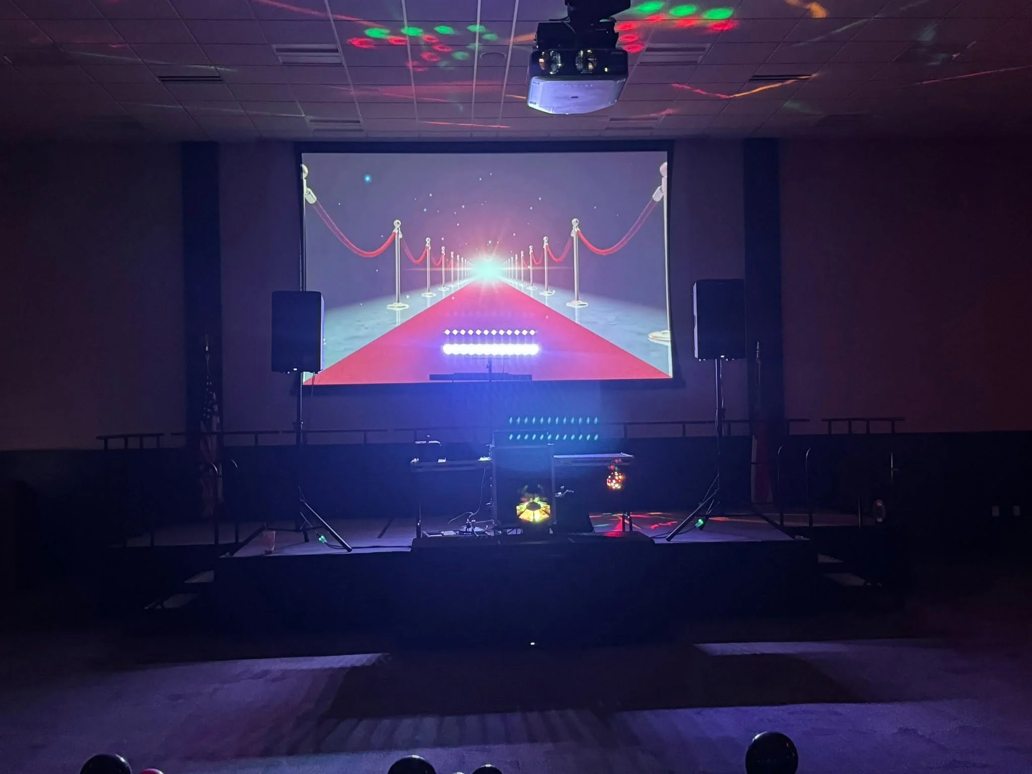 A dark room with a large screen displaying a virtual red carpet scene with velvet ropes, illuminated by colorful stage lights.