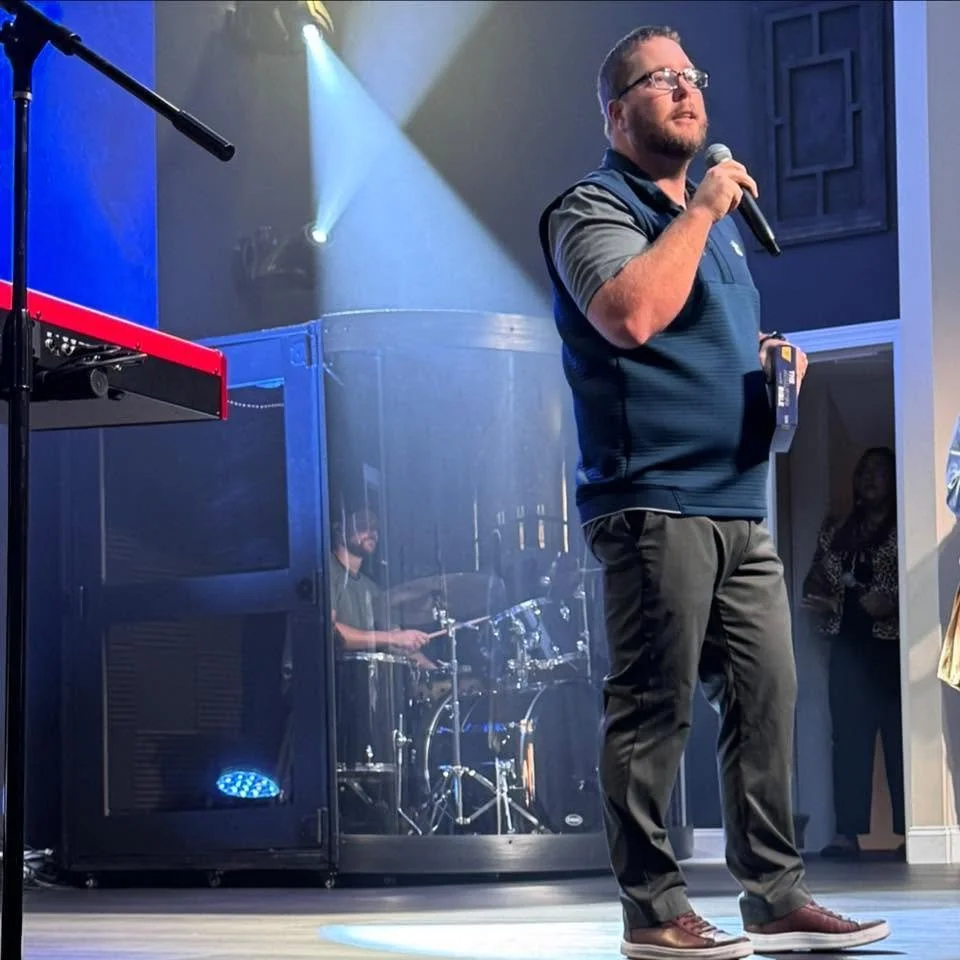Man holding microphone and standing on a stage with music equipment, including a drum set behind him, and people in the background.