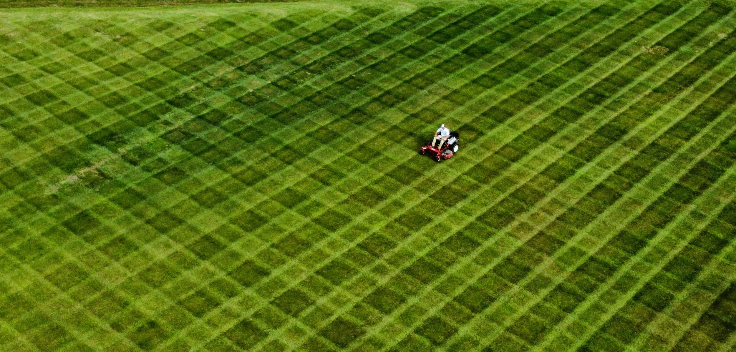 An aerial view of a tractor working on a large green field with neatly divided sections of grass or crops.