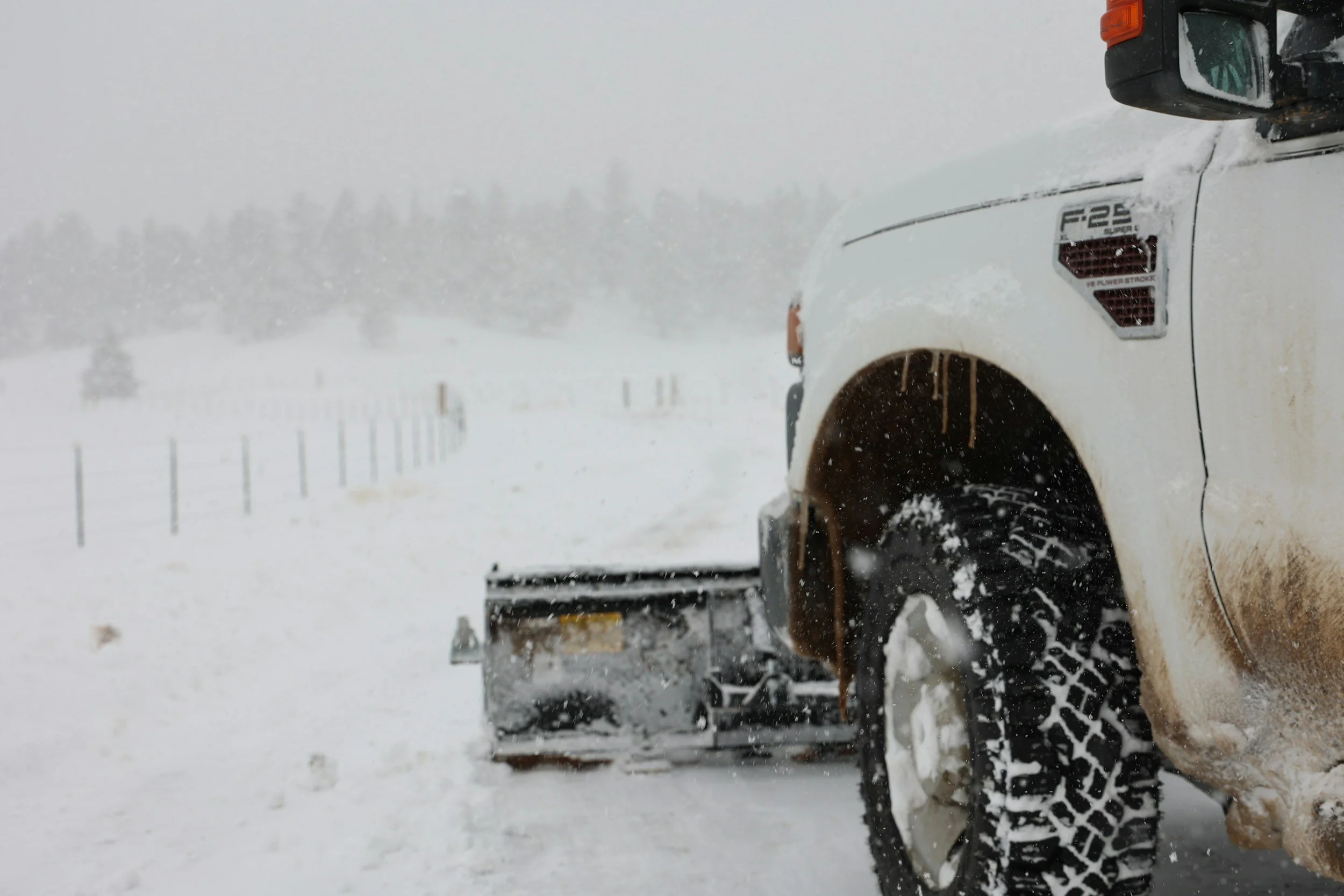 A white truck with a snowplow attachment clearing snow from a road during a snowstorm.