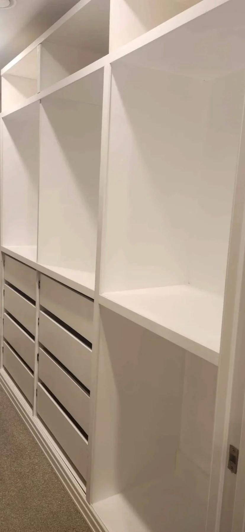 White custom-built shelving unit with open cubbies and six drawers on the bottom, installed in a corner of a room with beige walls and carpeted floor.