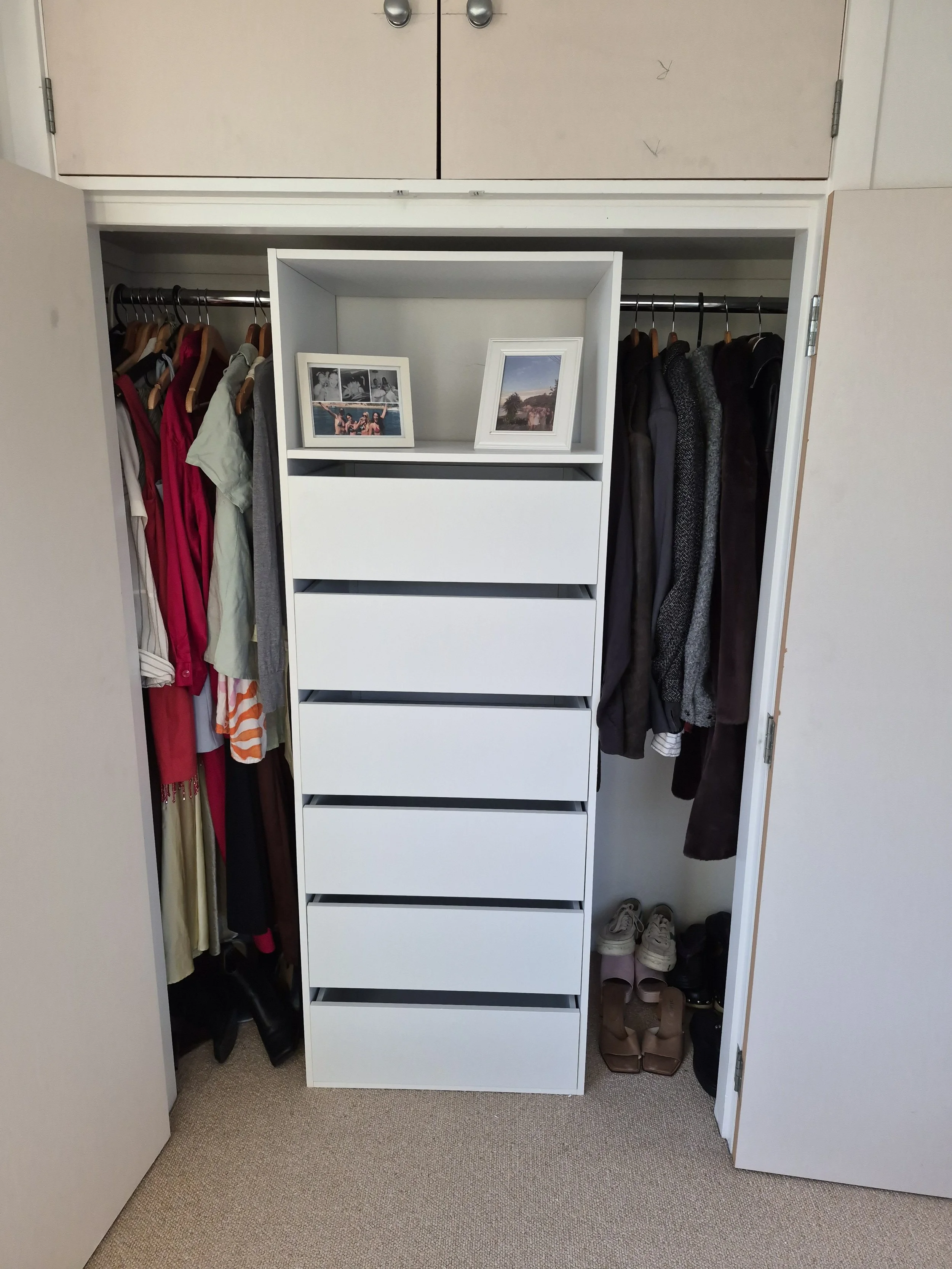 Open closet with hanging clothes on both sides, white drawers in the middle, framed photographs on top, shoes on the floor beneath, and closed upper cabinets.