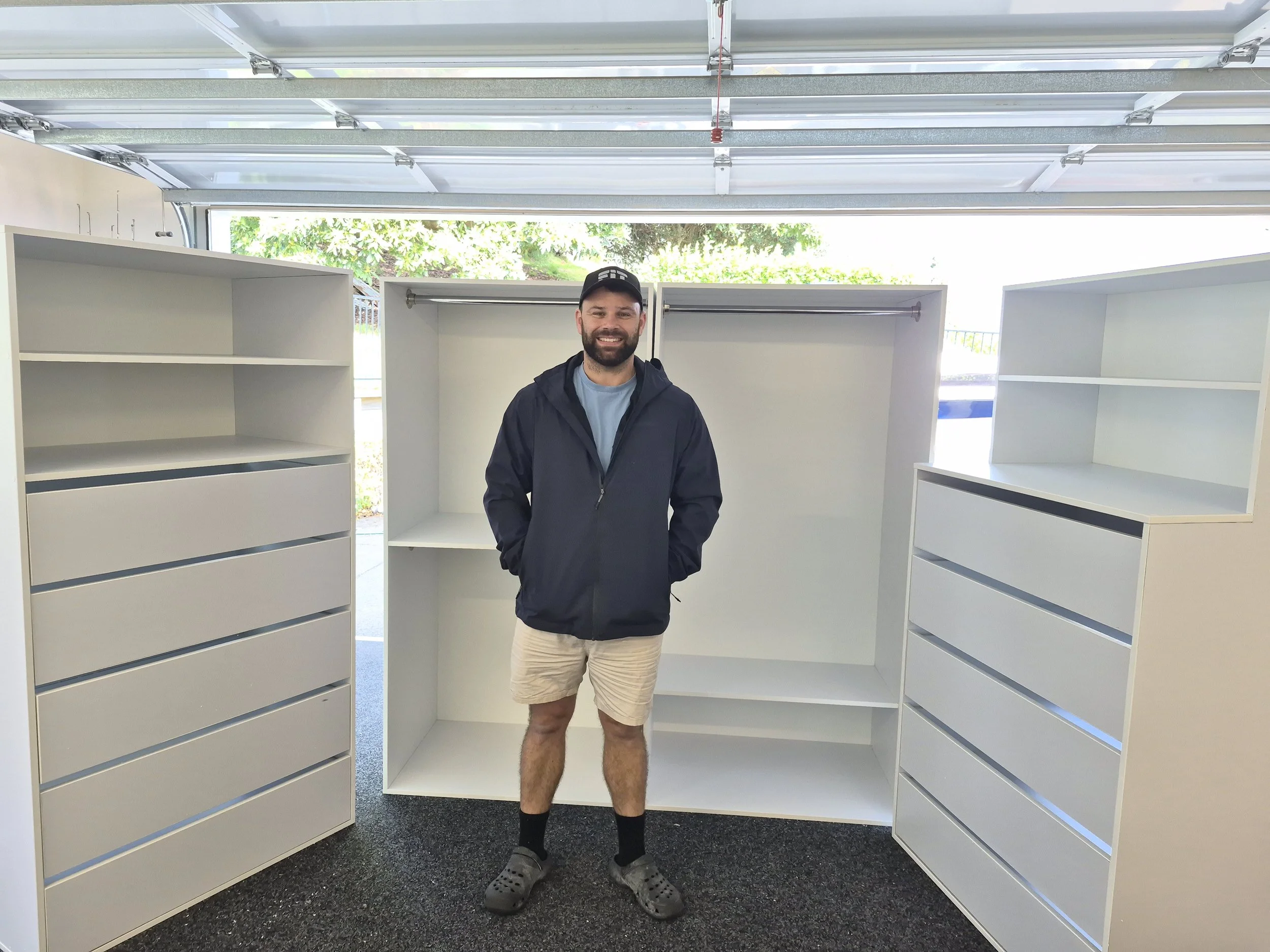 A smiling man with a beard and cap stands inside a wardrobe with empty white shelves and drawers, located in front of a garage door with windows showing greenery outside.